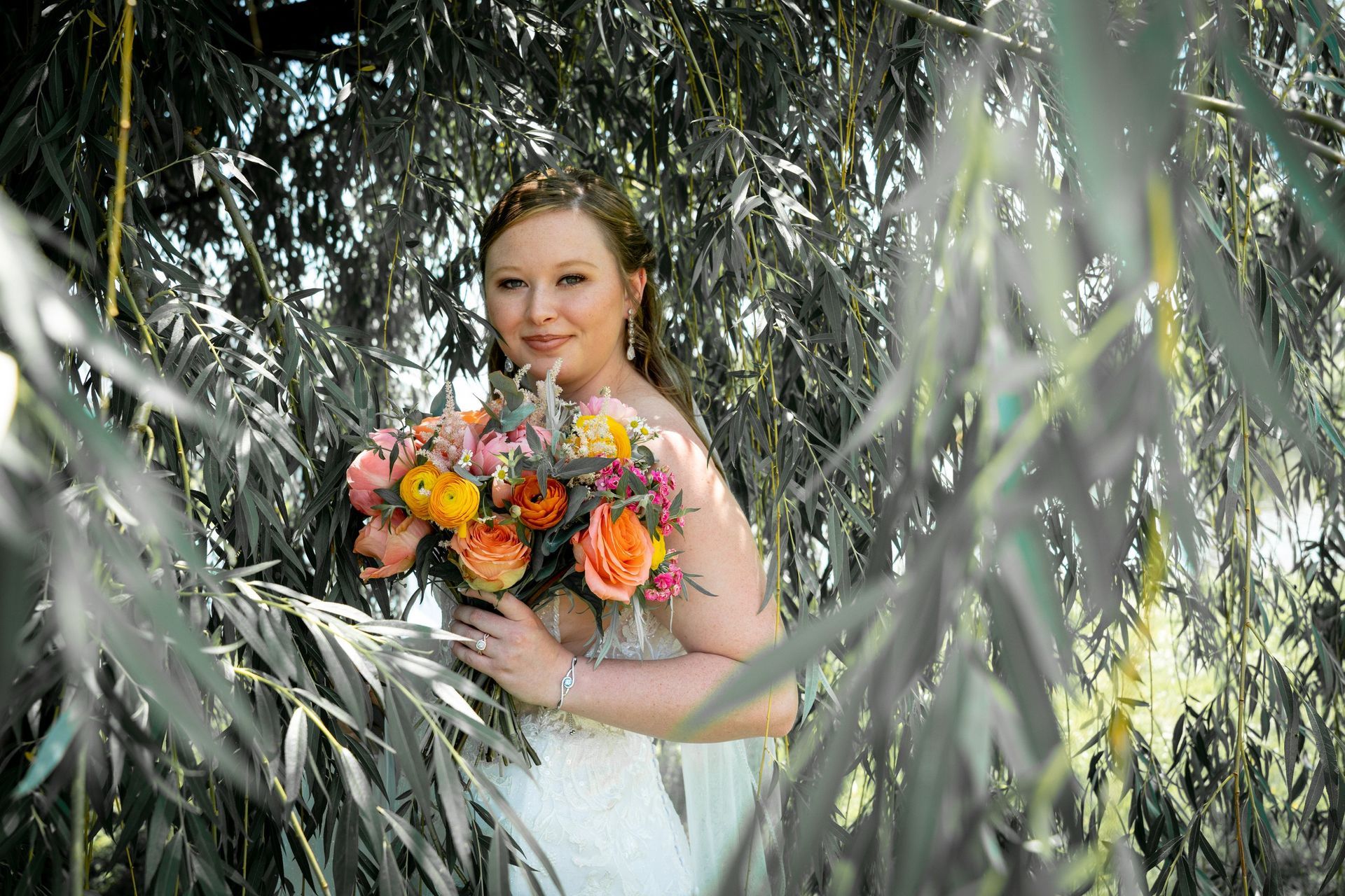 Bride with orange and pink bouquet poses under willow tree.