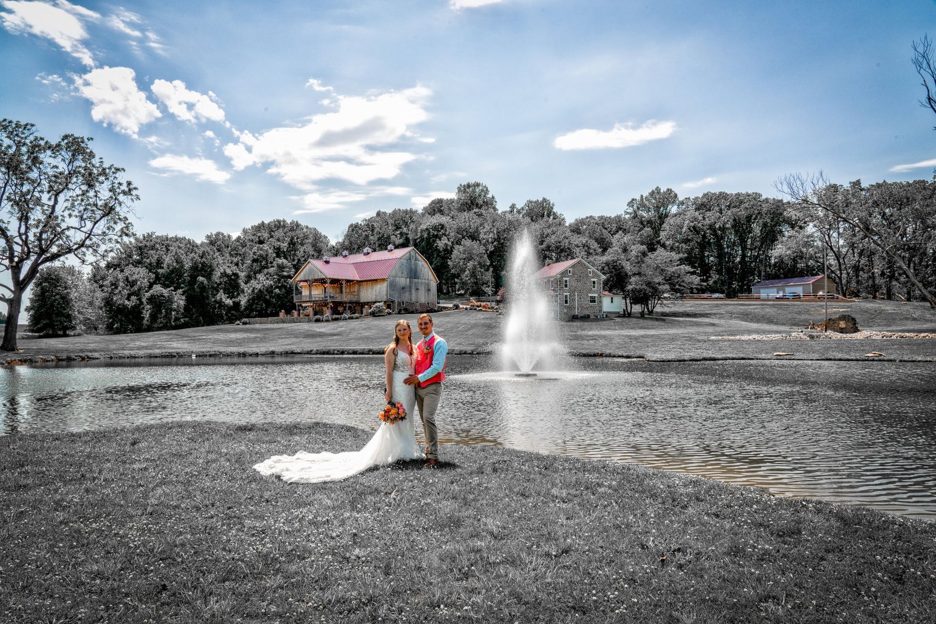 Bride and groom pose by pond with fountain; rural setting, muted colors except couple and sky.