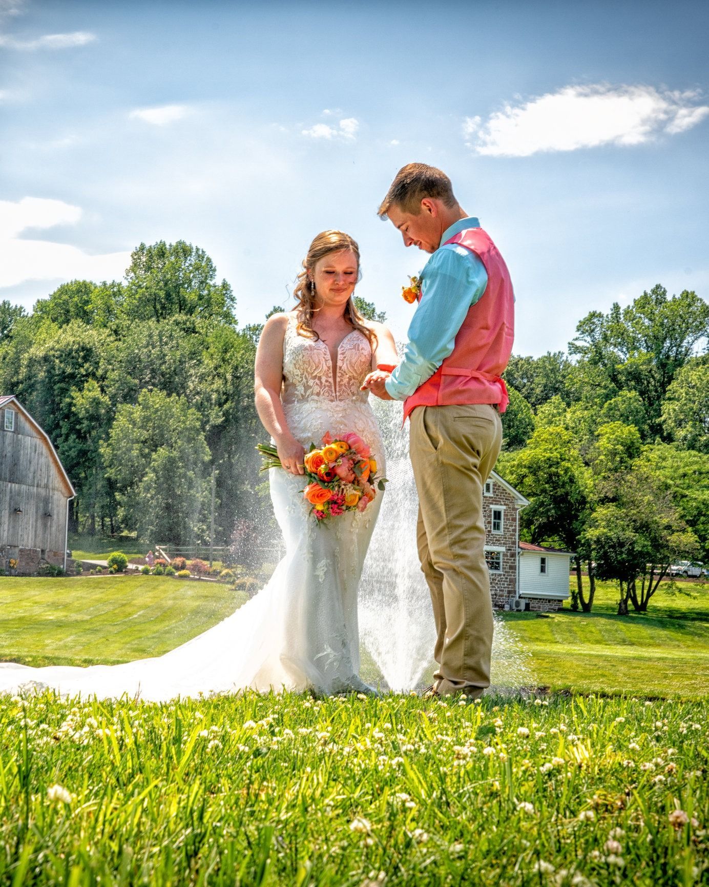 Bride and groom at outdoor wedding; she in white dress, he in pink vest, standing in field, blue sky.