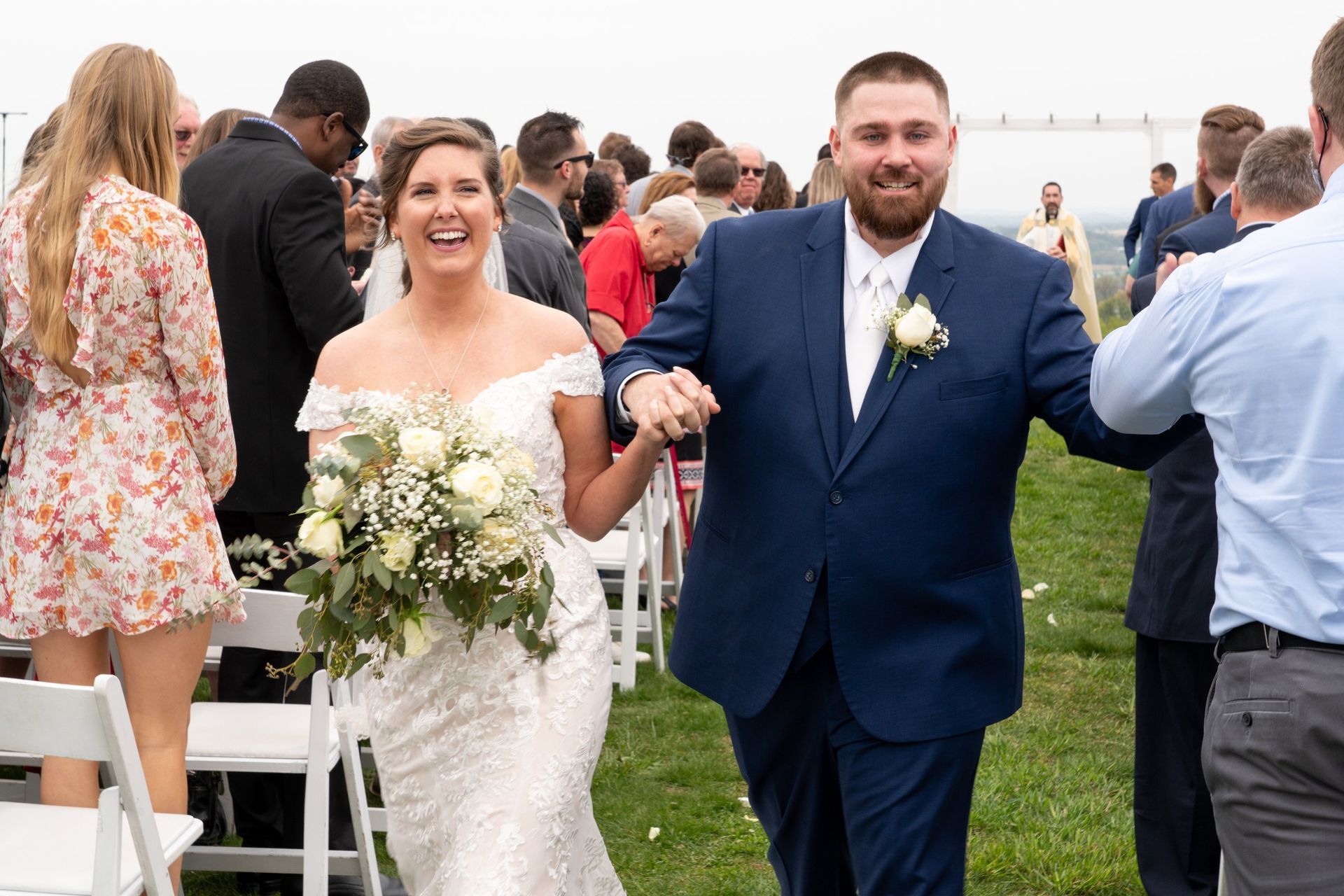 Newly married couple walking down aisle, smiling; woman in white dress, man in blue suit, outdoor wedding.