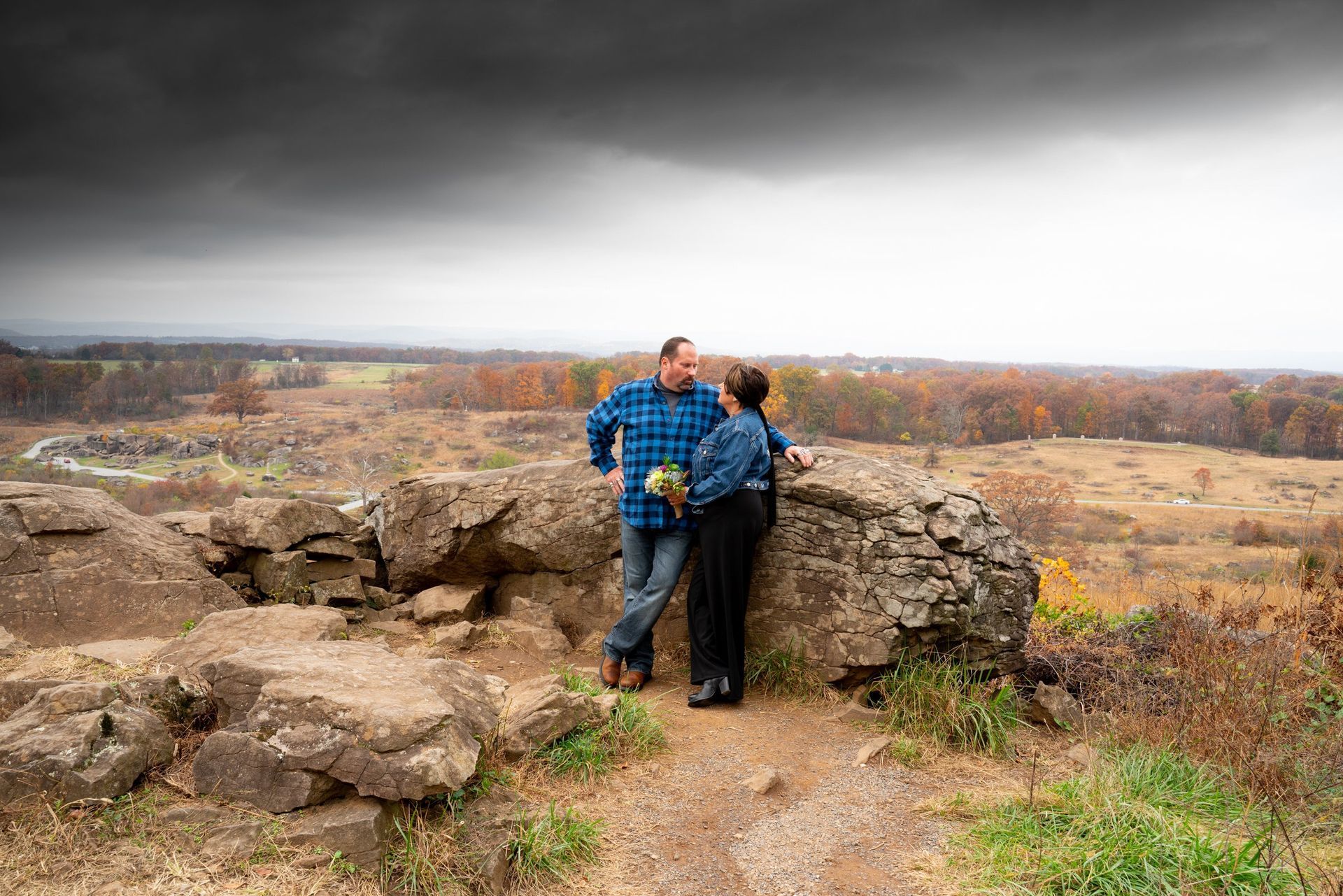Couple on a rocky overlook, gazing at a battlefield under a stormy sky.