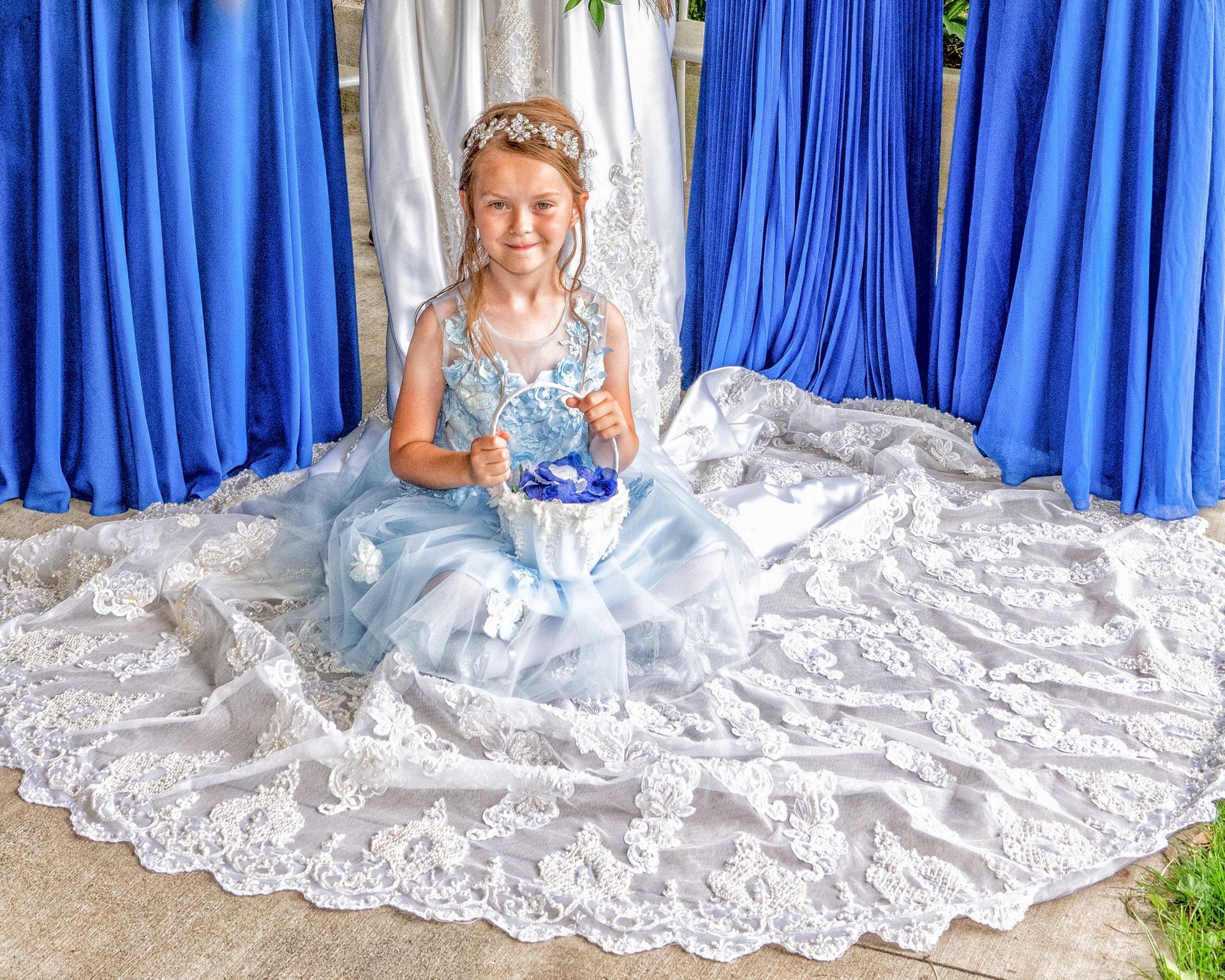 A young girl in a blue dress smiles, holding a basket, with bridesmaids in blue gowns behind her.