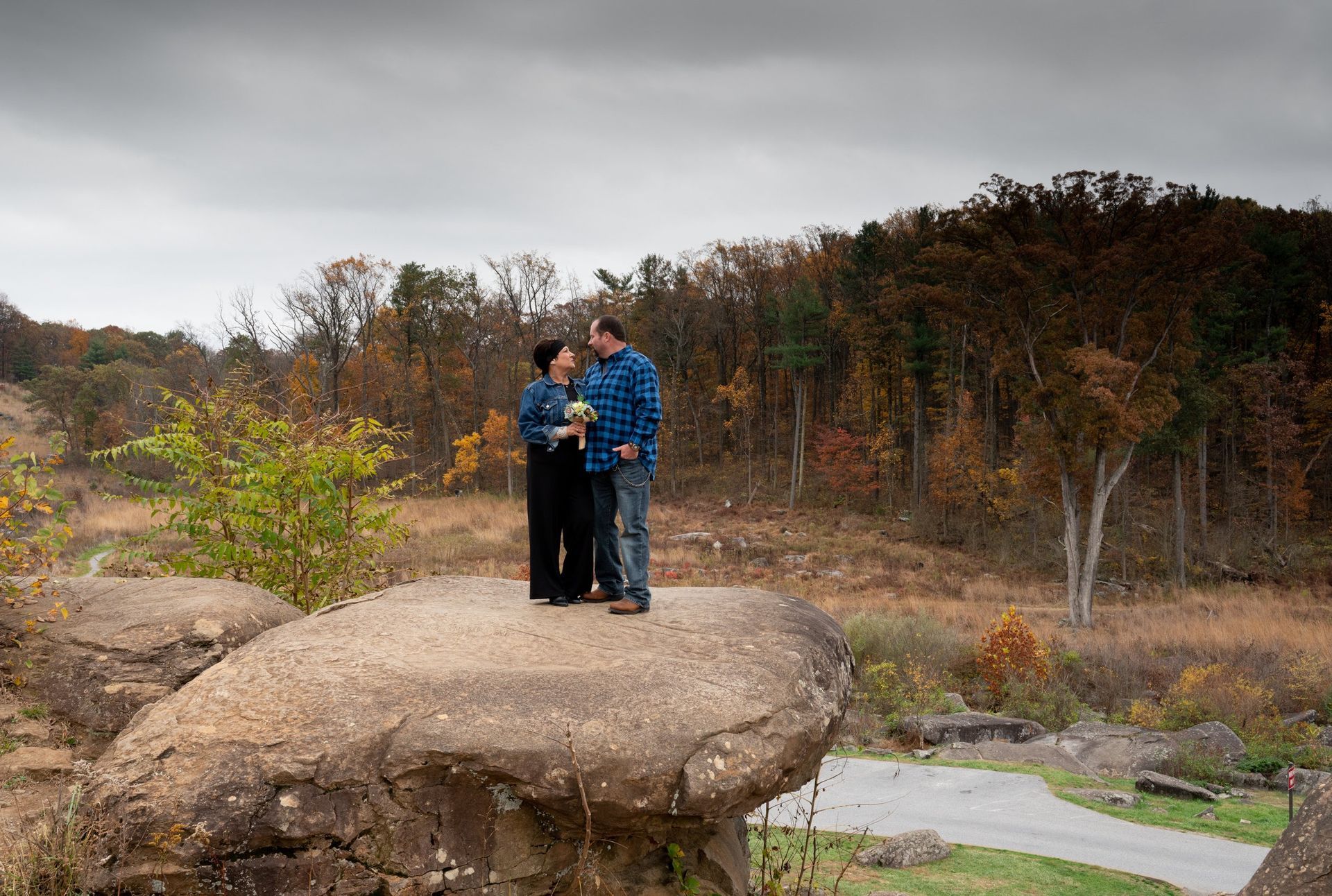 Couple stands on a large rock, holding each other, with fall foliage in the background.
