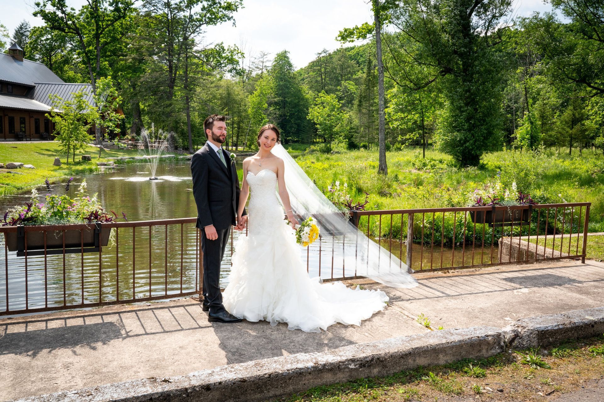 Bride and groom holding hands on a bridge, near a pond and trees, on a sunny day.