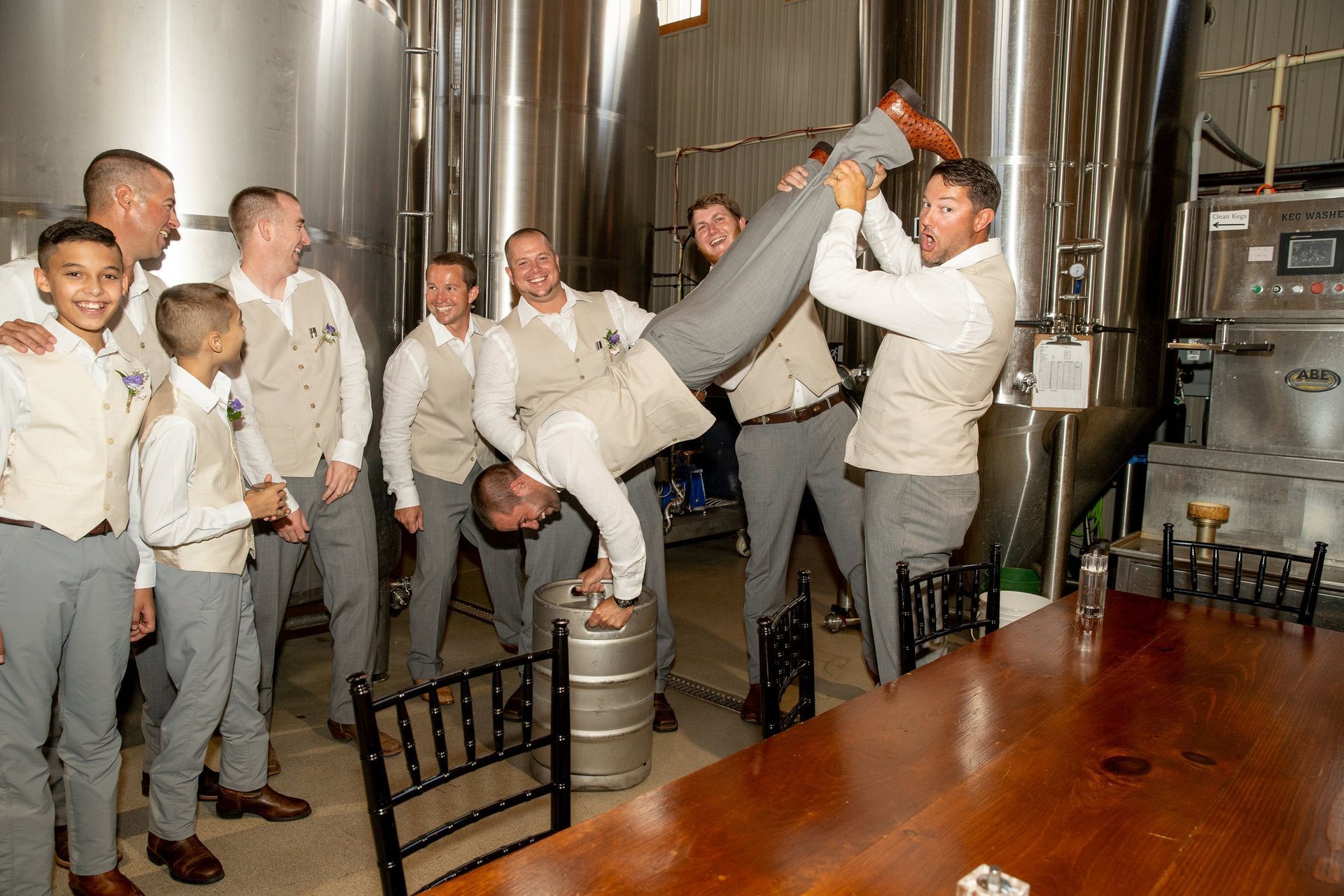 Groomsmen pose in brewery. Man doing handstand on keg as others watch and laugh; all wear vests and gray pants.