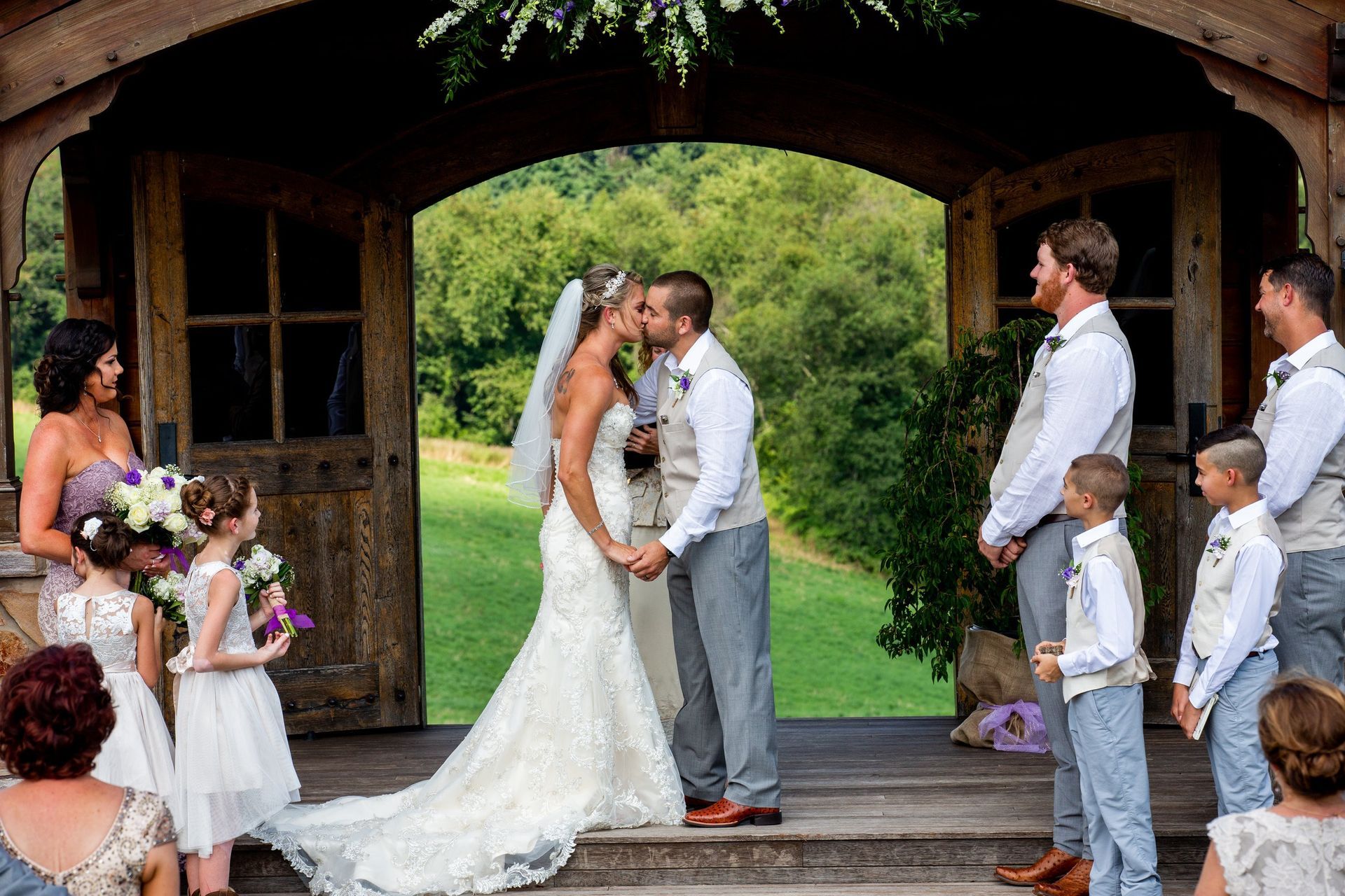 Bride and groom kiss at outdoor wedding, surrounded by wedding party and backdrop of green hills.