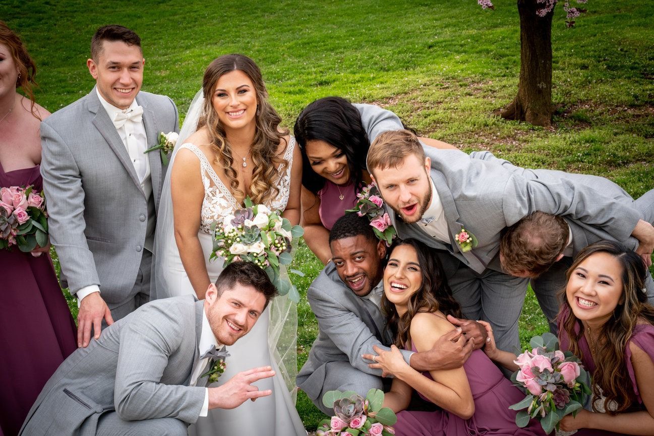 Wedding party poses playfully on a grassy lawn. Bride and groom with bridesmaids and groomsmen smiling and laughing.