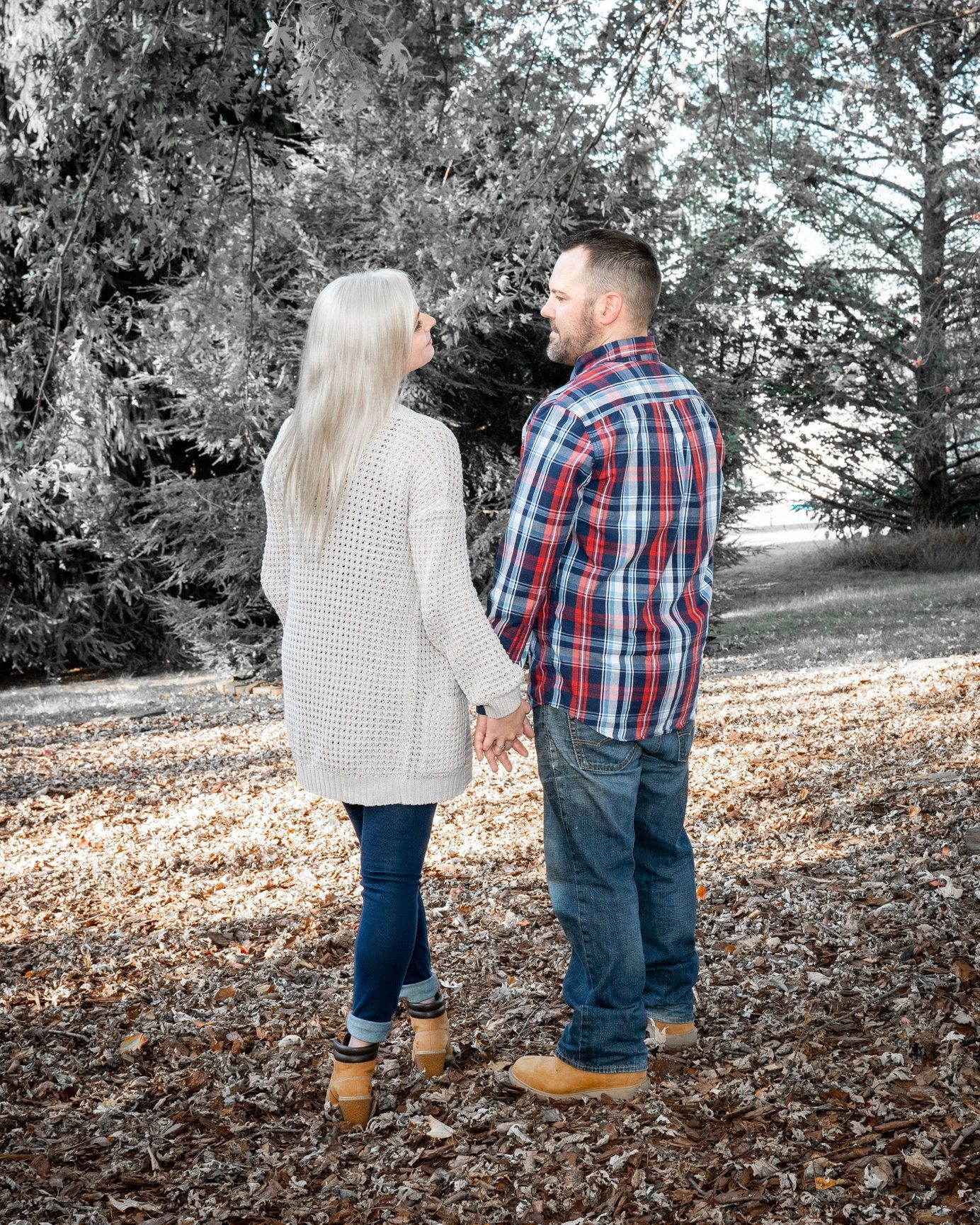 Couple holding hands, facing away, standing on leaves. Woman blonde, wearing white sweater. Man wearing plaid shirt, jeans.