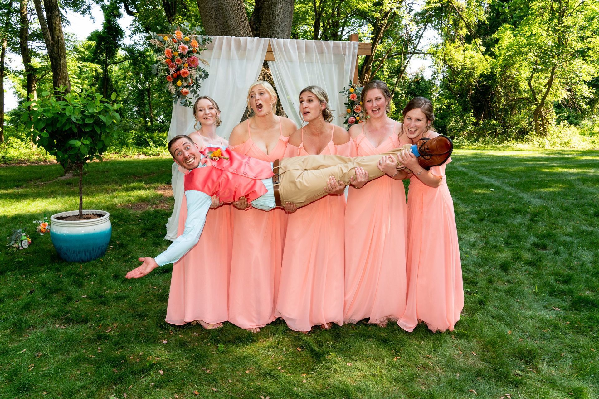 Bridesmaids in peach dresses lifting a smiling man in front of a floral backdrop on a lawn.