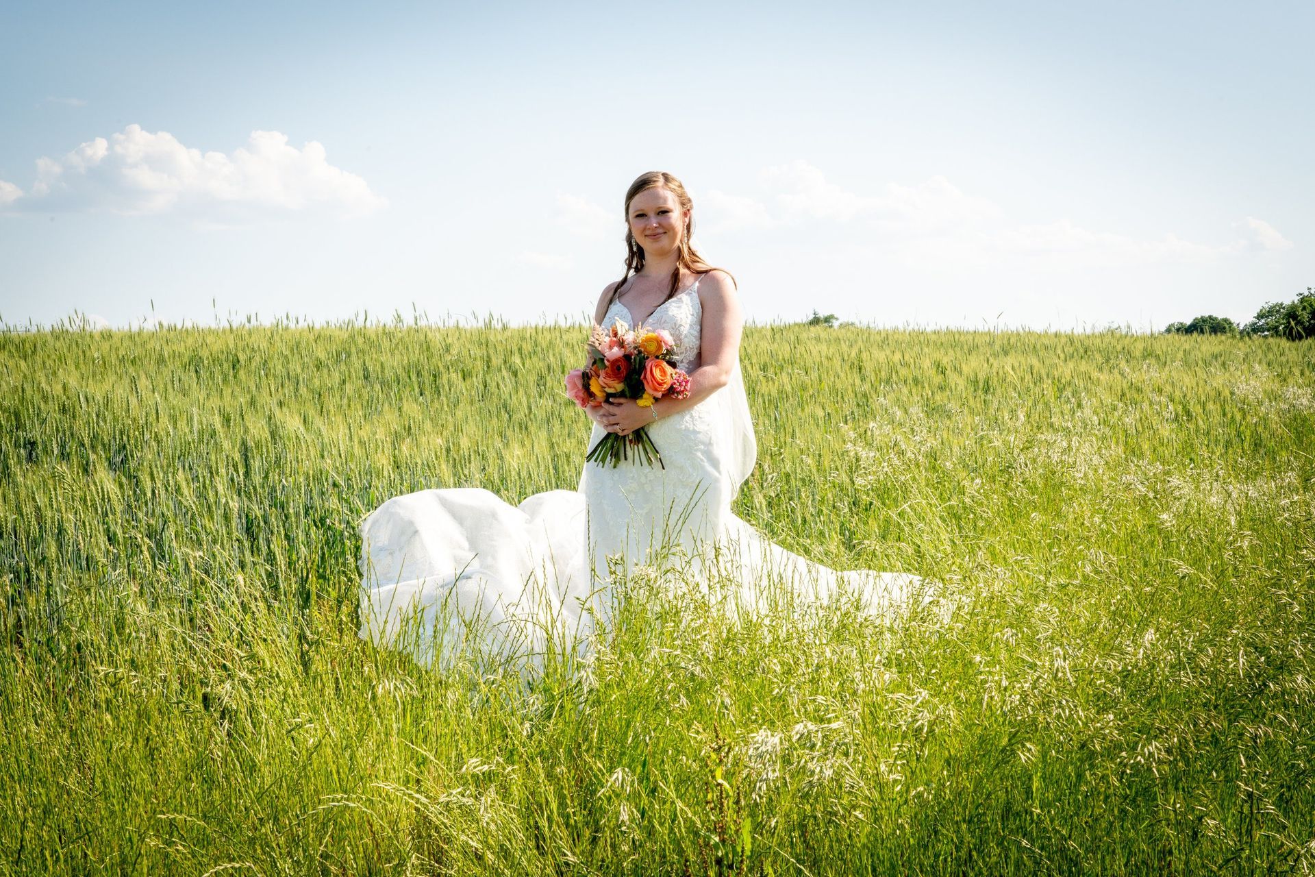 Bride in white dress holding flowers, standing in a green field on a sunny day.