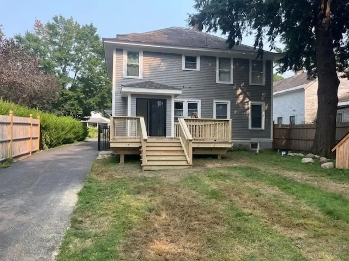 Gray house with wooden deck and stairs; grassy lawn and driveway.