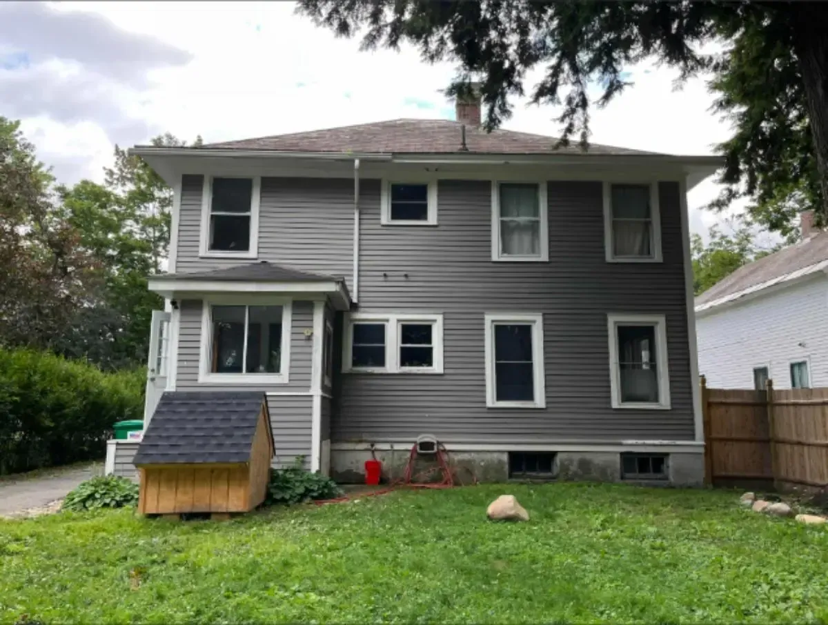 Two-story gray house with wooden deck and steps. Front yard with dry grass and driveway.