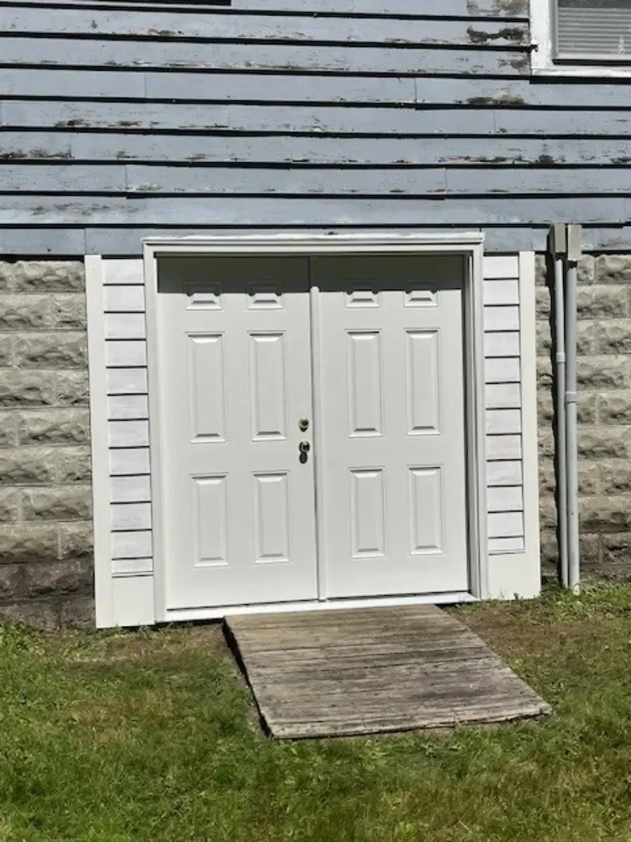 White double doors with a wooden ramp leading up, set in a weathered blue building exterior with stone foundation.