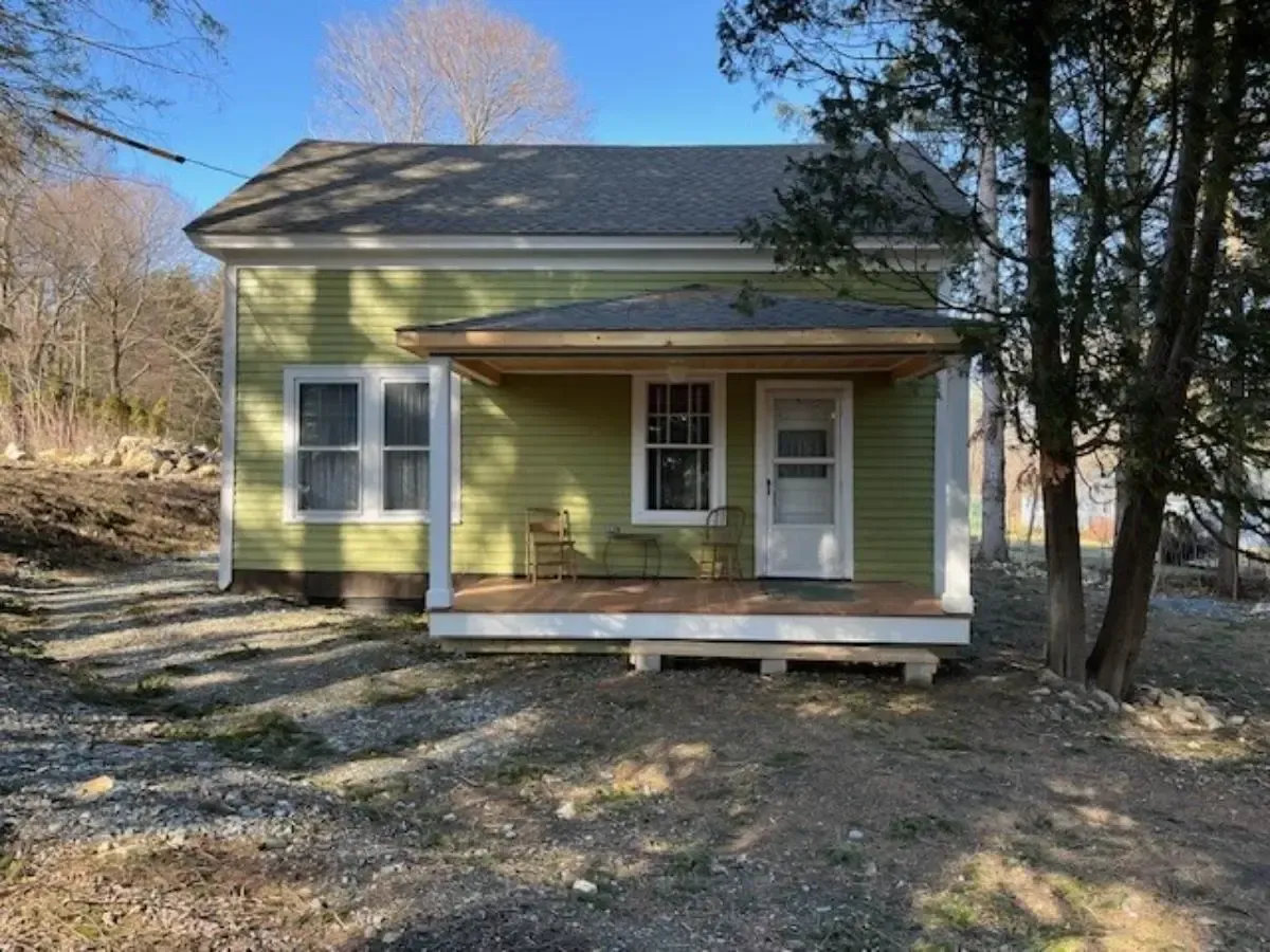 A small green house with a porch and trees in the background.