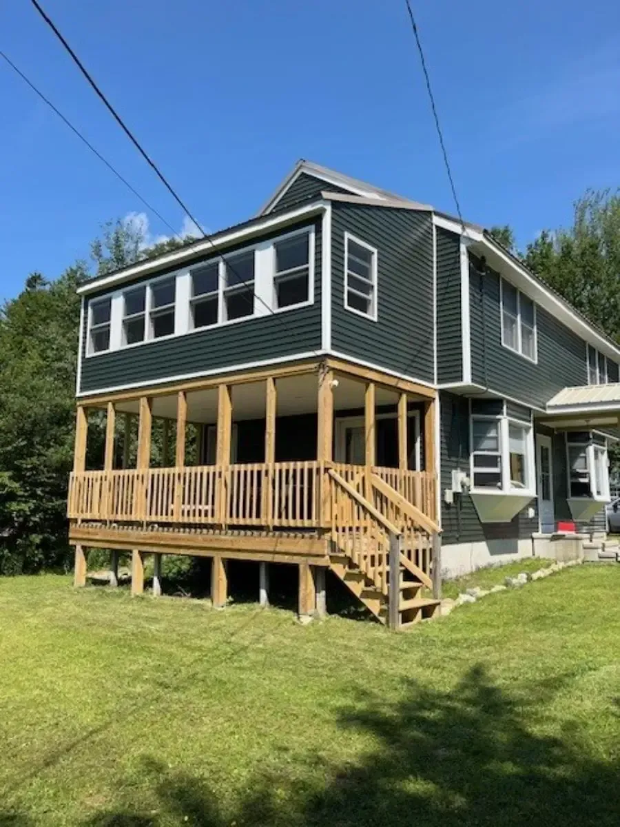 Two-story house with green siding, wooden porch, and stairs on a grassy lawn.