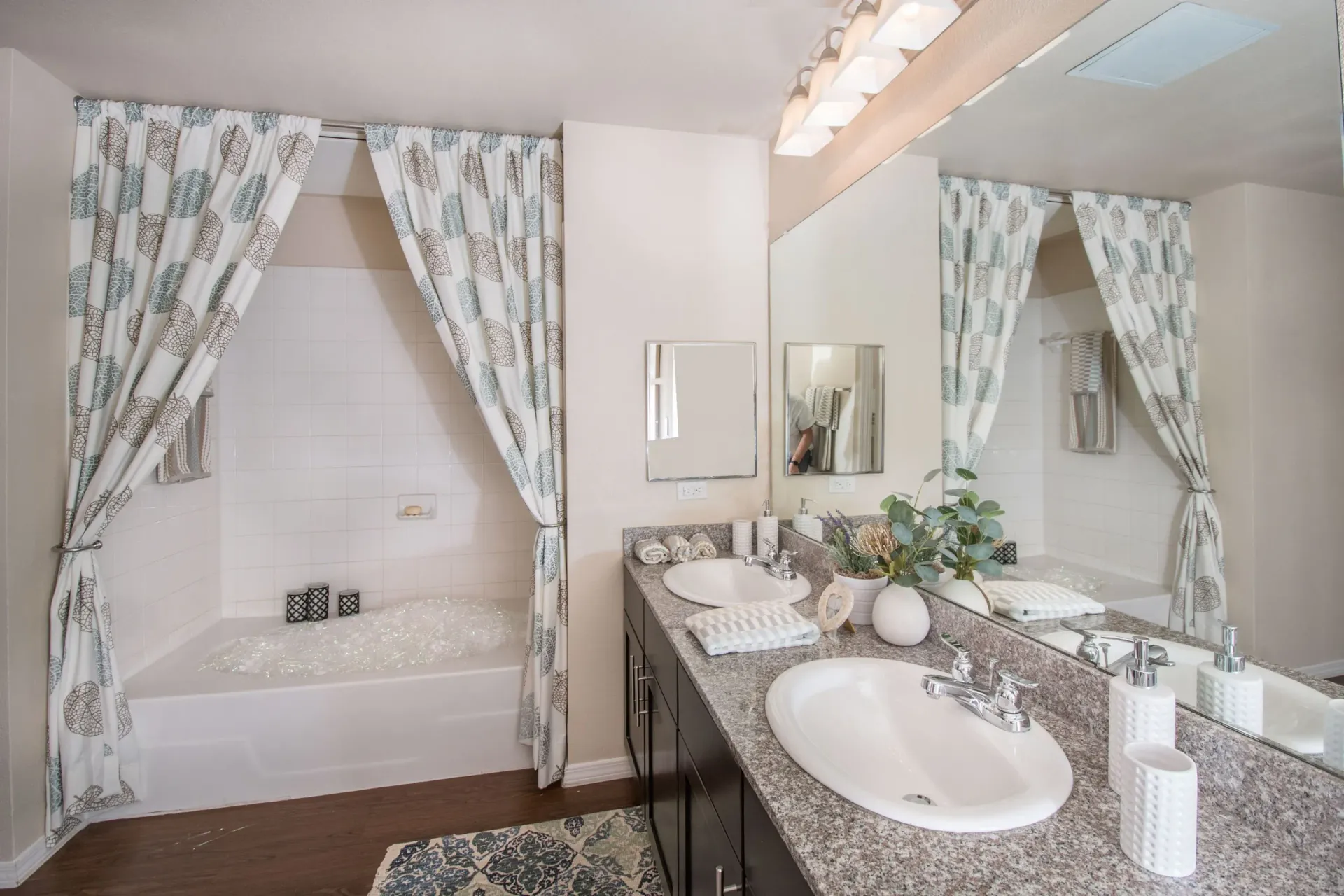 Bathroom with double-sink vanity, granite countertop, mirrors, and a tiled tub with patterned curtains.