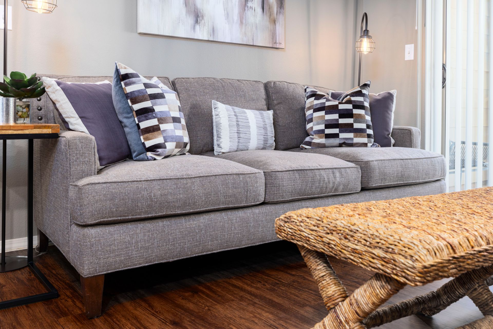 Gray sofa with patterned throw pillows in a room with wood flooring, a woven ottoman, and a side table with a plant.