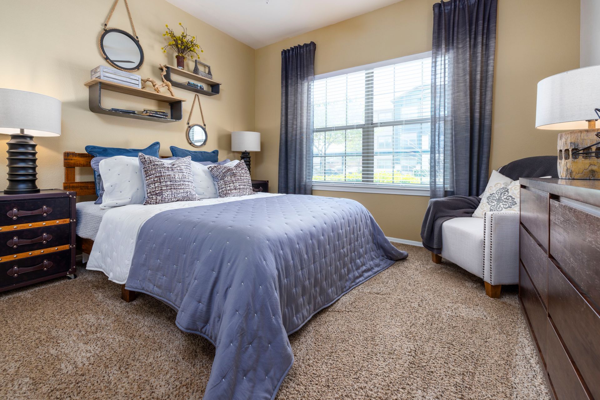 A bedroom featuring a queen bed with blue bedding, a side chair, window curtains, and decorative wall shelves.
