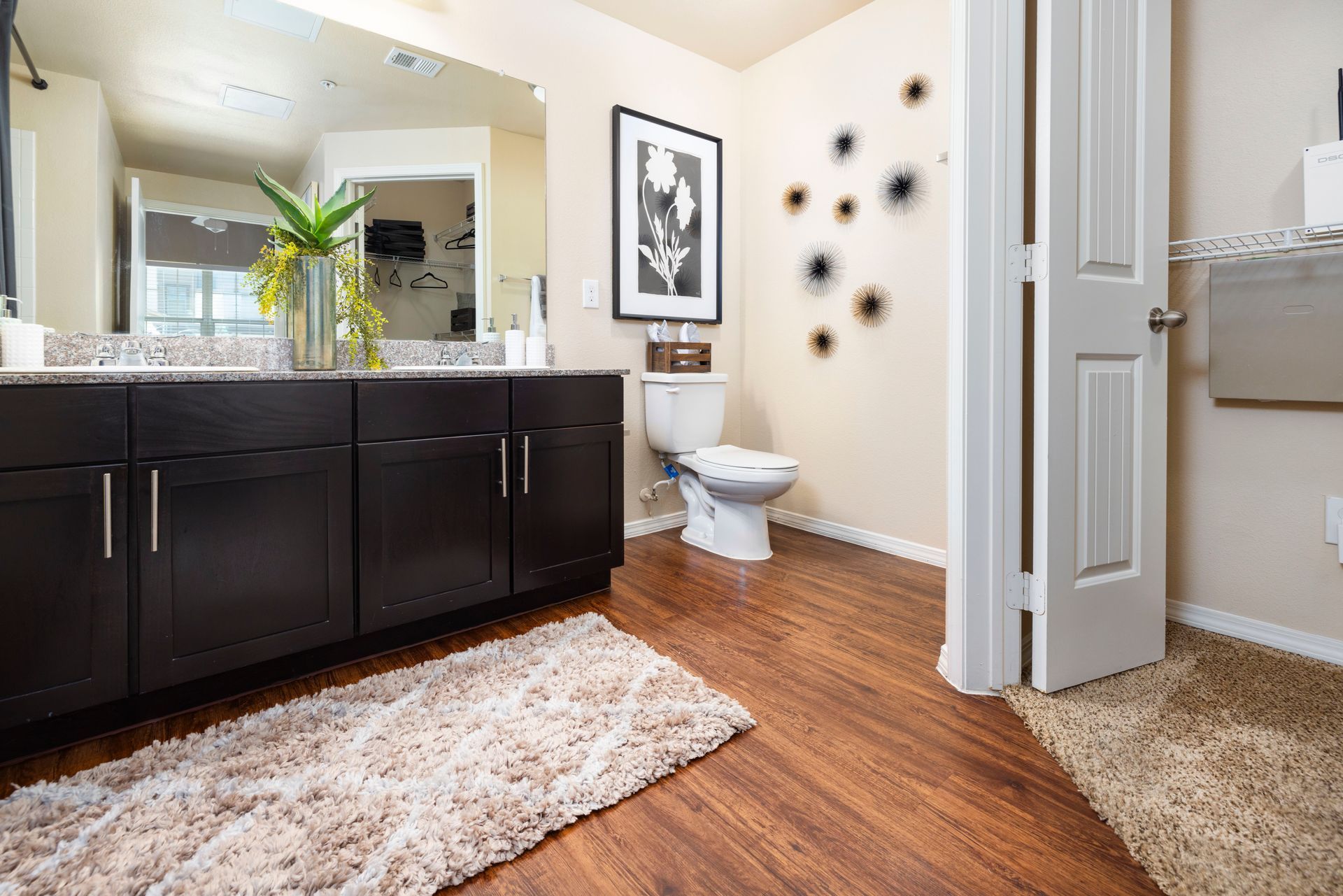 A bathroom with dark brown cabinets, a large mirror, a white toilet, wood-look flooring, and a cream-colored rug.