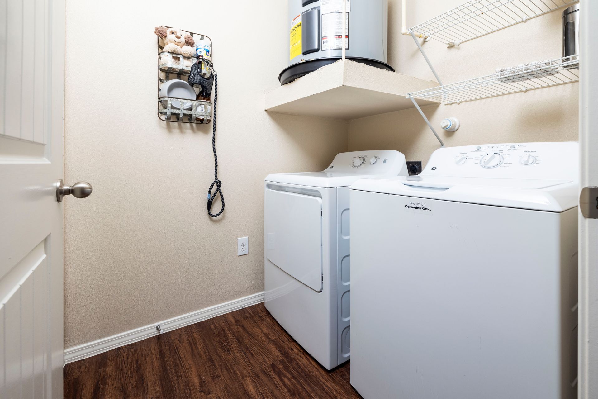 A laundry room with white appliances, a water heater on an overhead shelf, wood flooring, and a wall-mounted organizer.