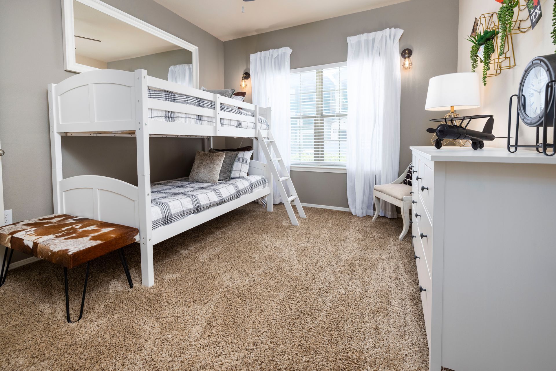 A bedroom features a white bunk bed, a cowhide-patterned bench, a white dresser, and white curtains by a window.