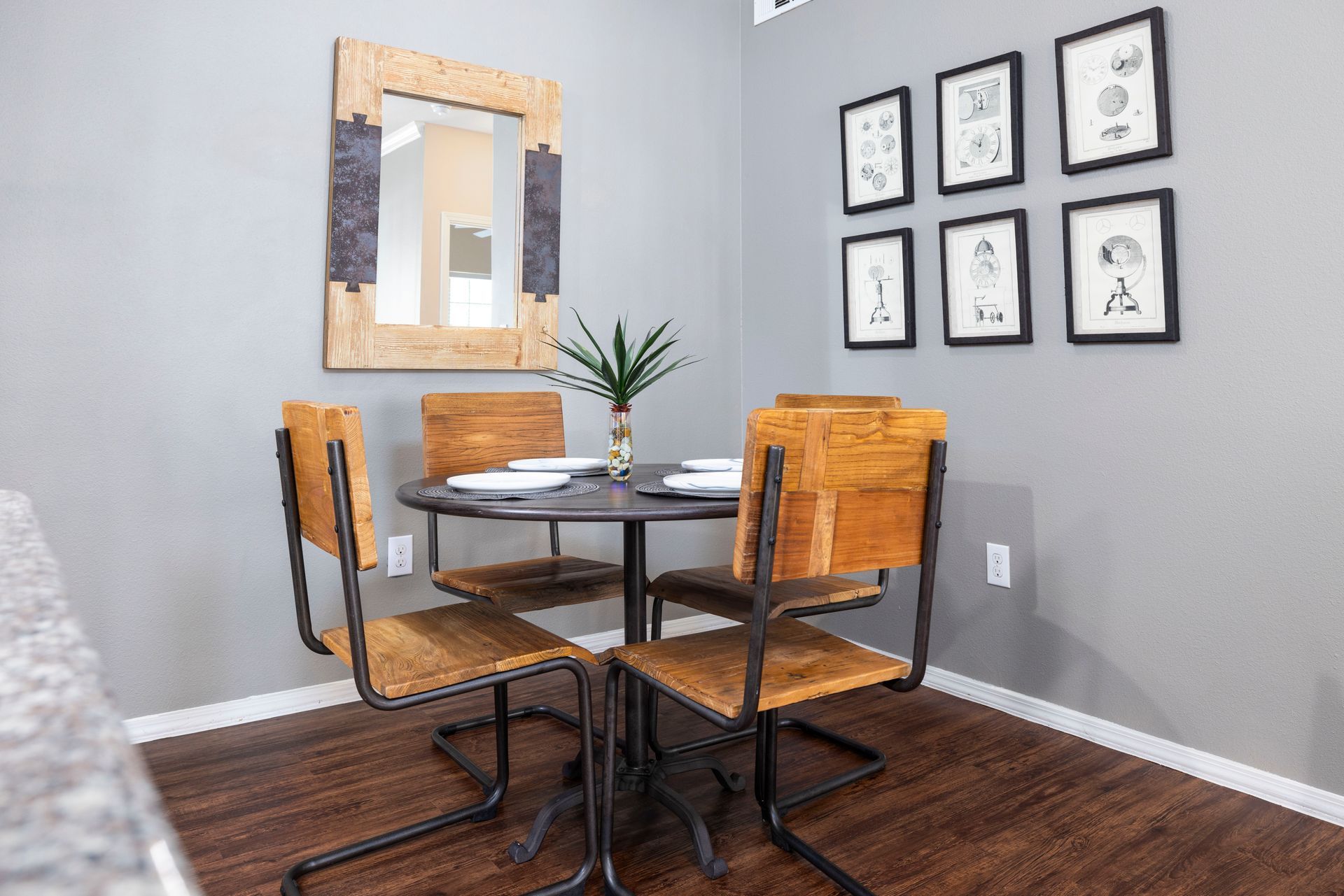 A dining set with a round table and four wooden chairs sits in a room with a large wooden mirror and six wall art frames.