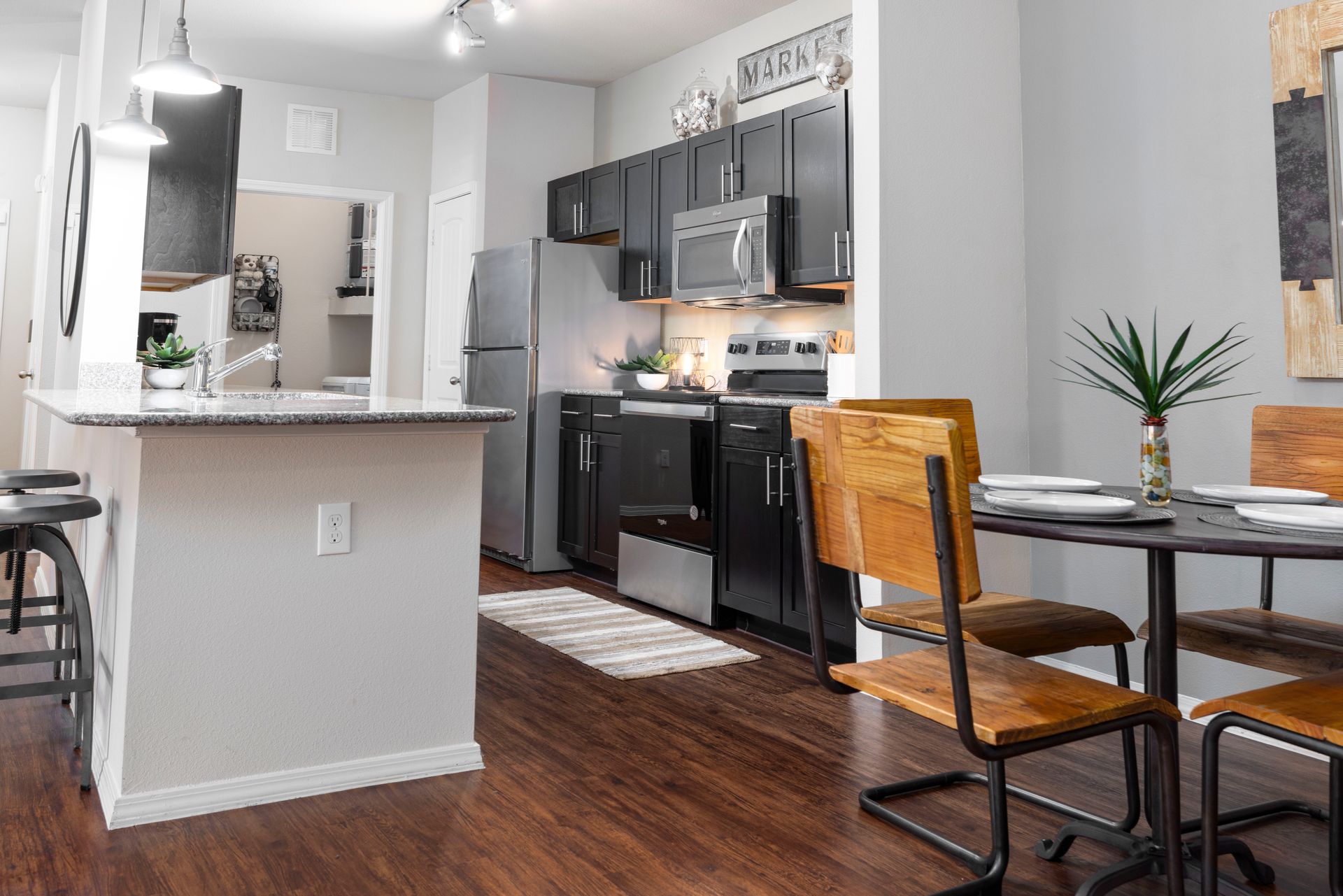 A modern apartment kitchen with dark cabinets, stainless steel appliances, an island, and a small wooden dining set.