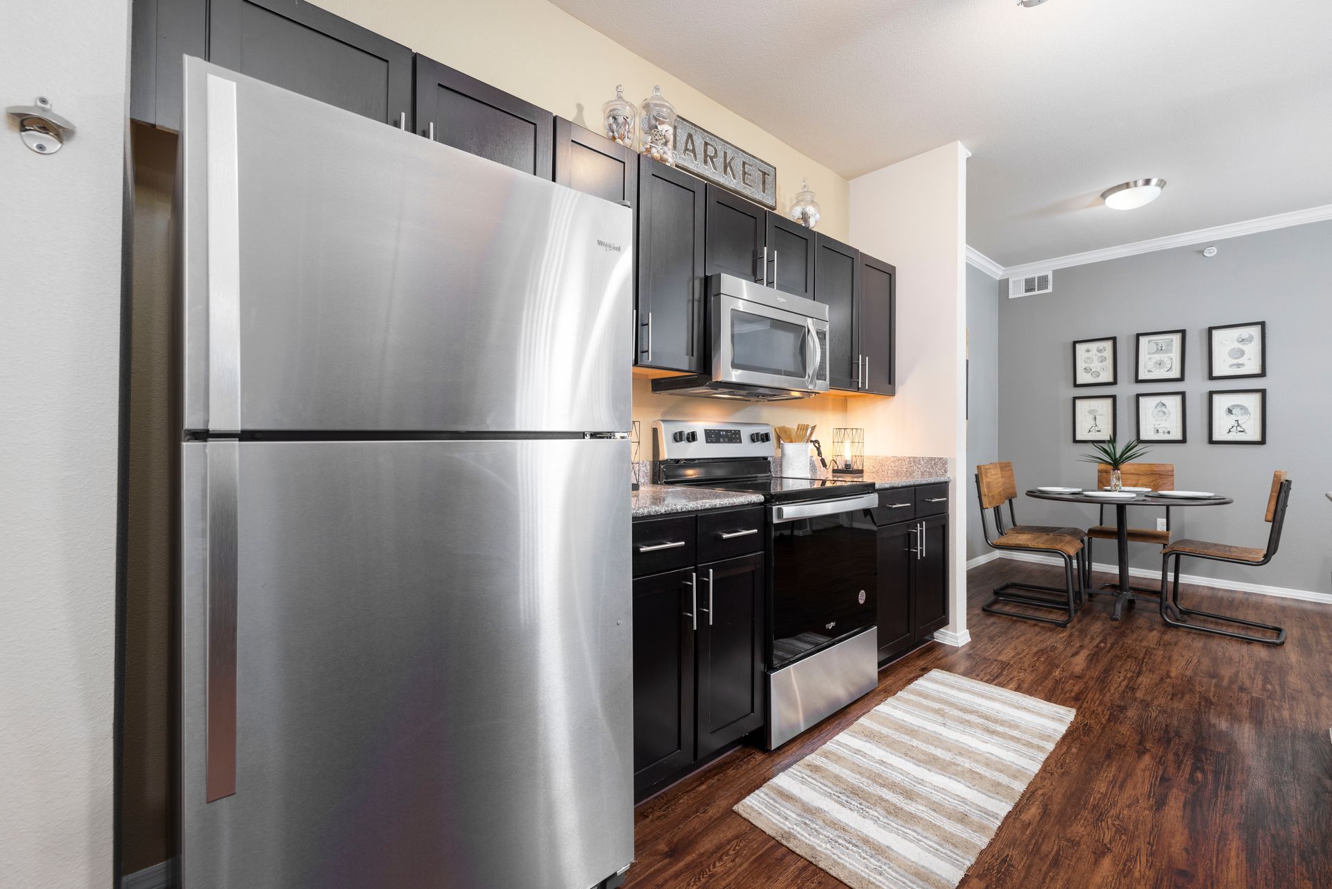 Modern kitchen with stainless steel appliances, dark cabinets, and a small dining area with four framed wall prints.
