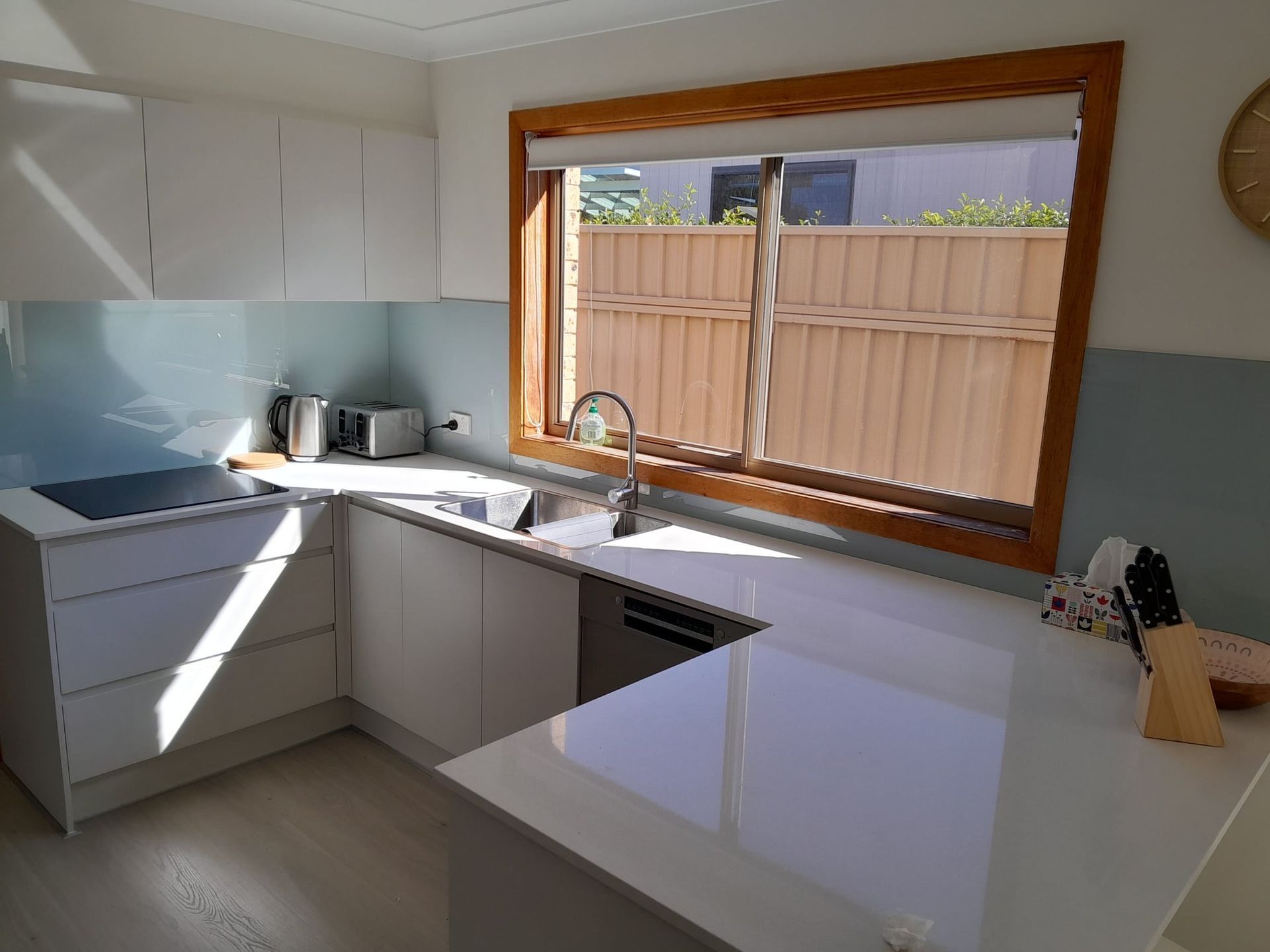 Modern White Kitchen With Light Blue Backsplash And Window Overlooking A Wooden Fence
