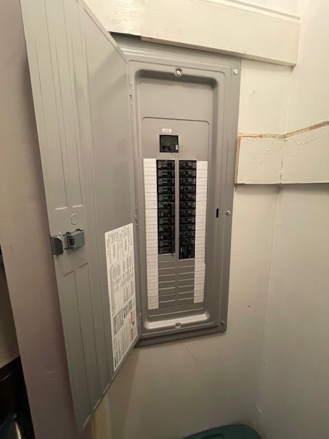 Opened gray electrical panel with rows of black and white circuit breakers, mounted on a wall.