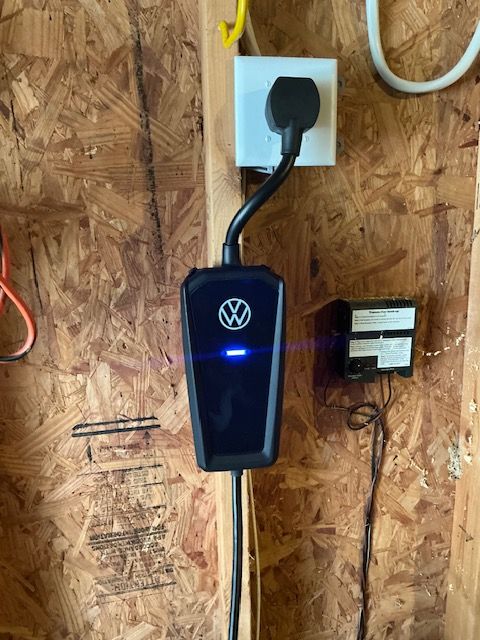 Volkswagen EV charger with blue light, plugged into a wall outlet on a wood wall.