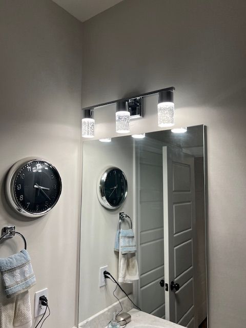 Bathroom with mirror, lights, clock, and towel. Grey walls, white door, black accents.