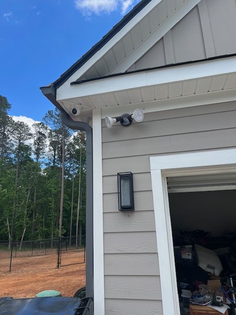Exterior of a building with security cameras and lights, light gray siding, white trim, and a blue sky.