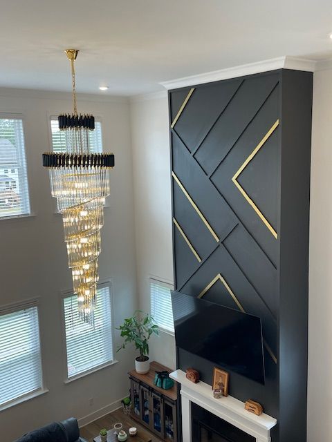 Modern living room: black accent wall with gold trim, large chandelier, TV, fireplace, and windows.