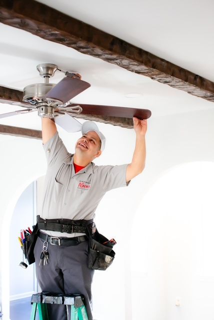 Electrician installing a ceiling fan; he's smiling, wearing work clothes and a tool belt; interior setting.