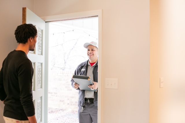 Man answers door to greet service worker holding a clipboard, in a bright doorway.