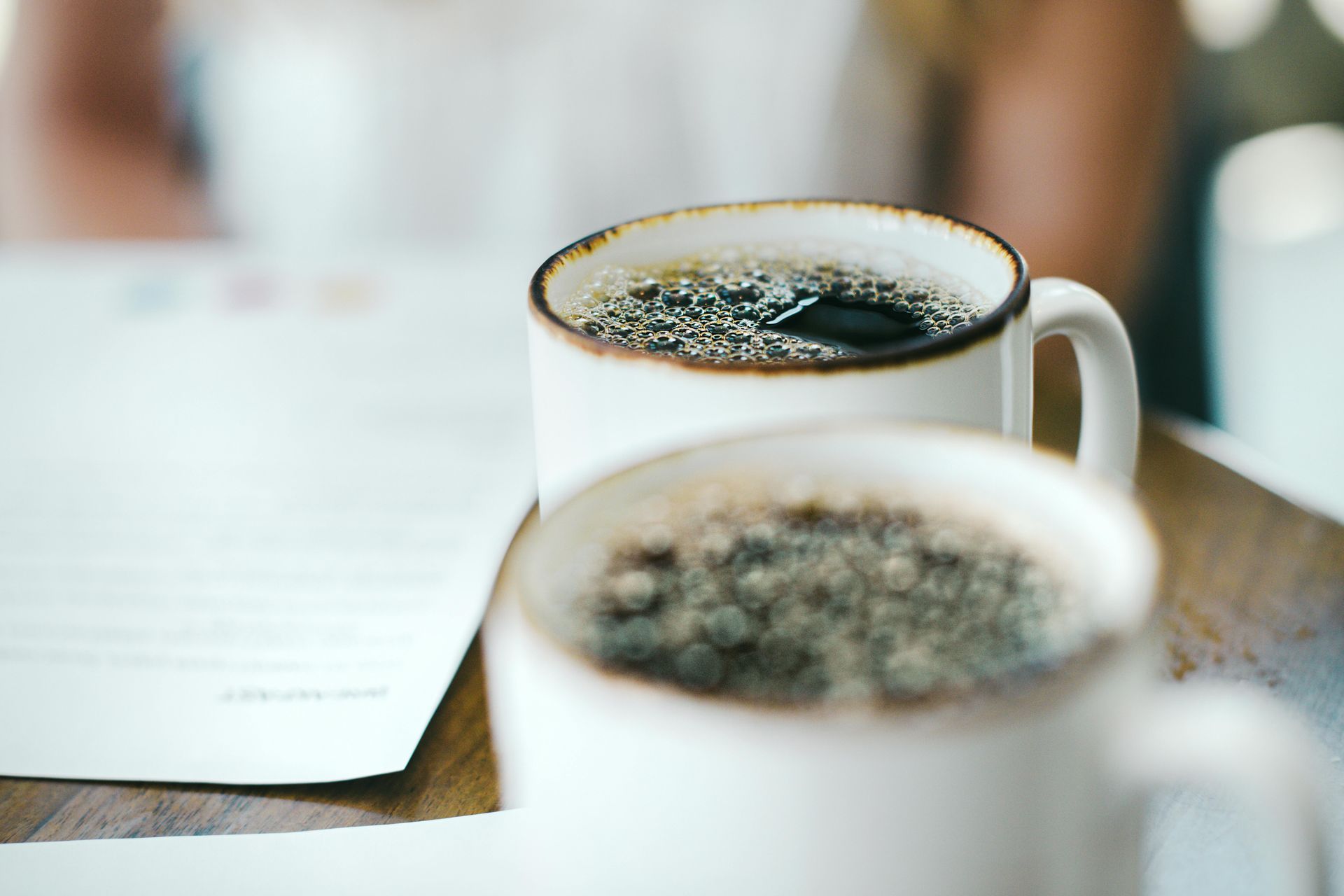 Two white mugs of coffee on a wooden table, with a blurred background of a person and papers.