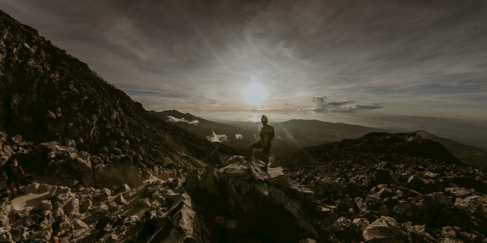 Person atop rocky mountain, facing sunlit horizon. Landscape view with cloudy sky.