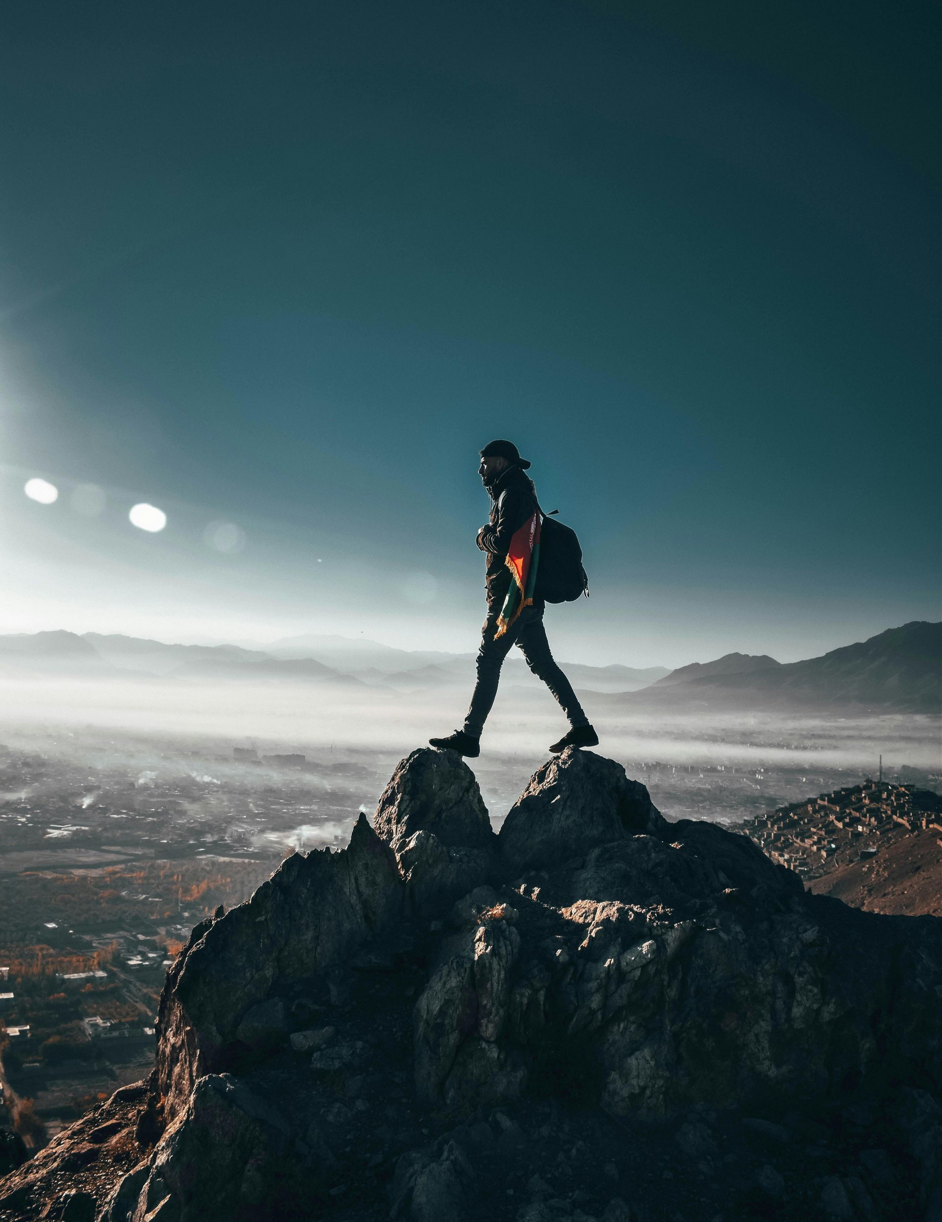 Person hiking on mountain peak, backpack. City and mist in background. Blue sky, sunlight.