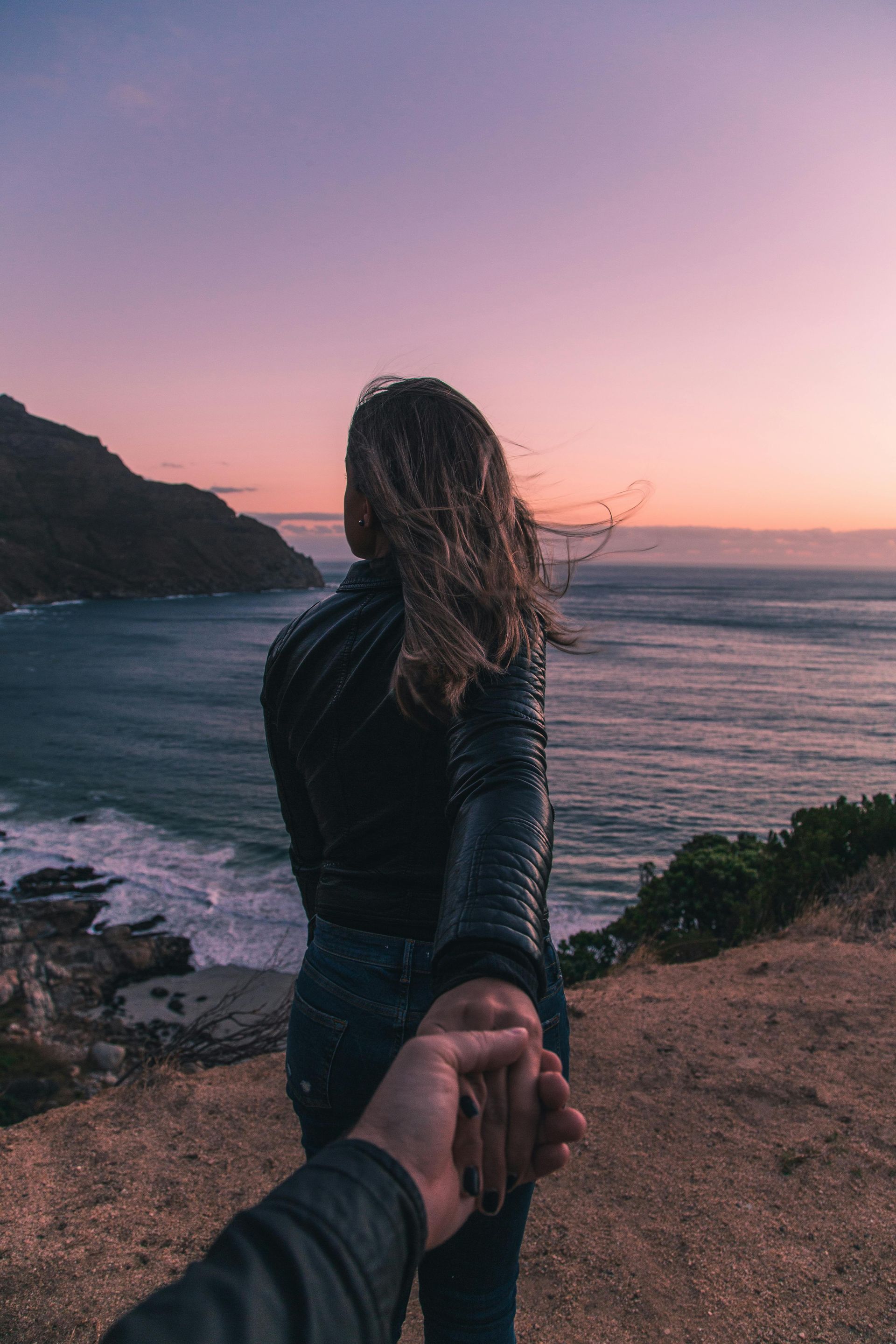 Woman leading a hand towards ocean at sunset; coast and mountains in background, holding hands.