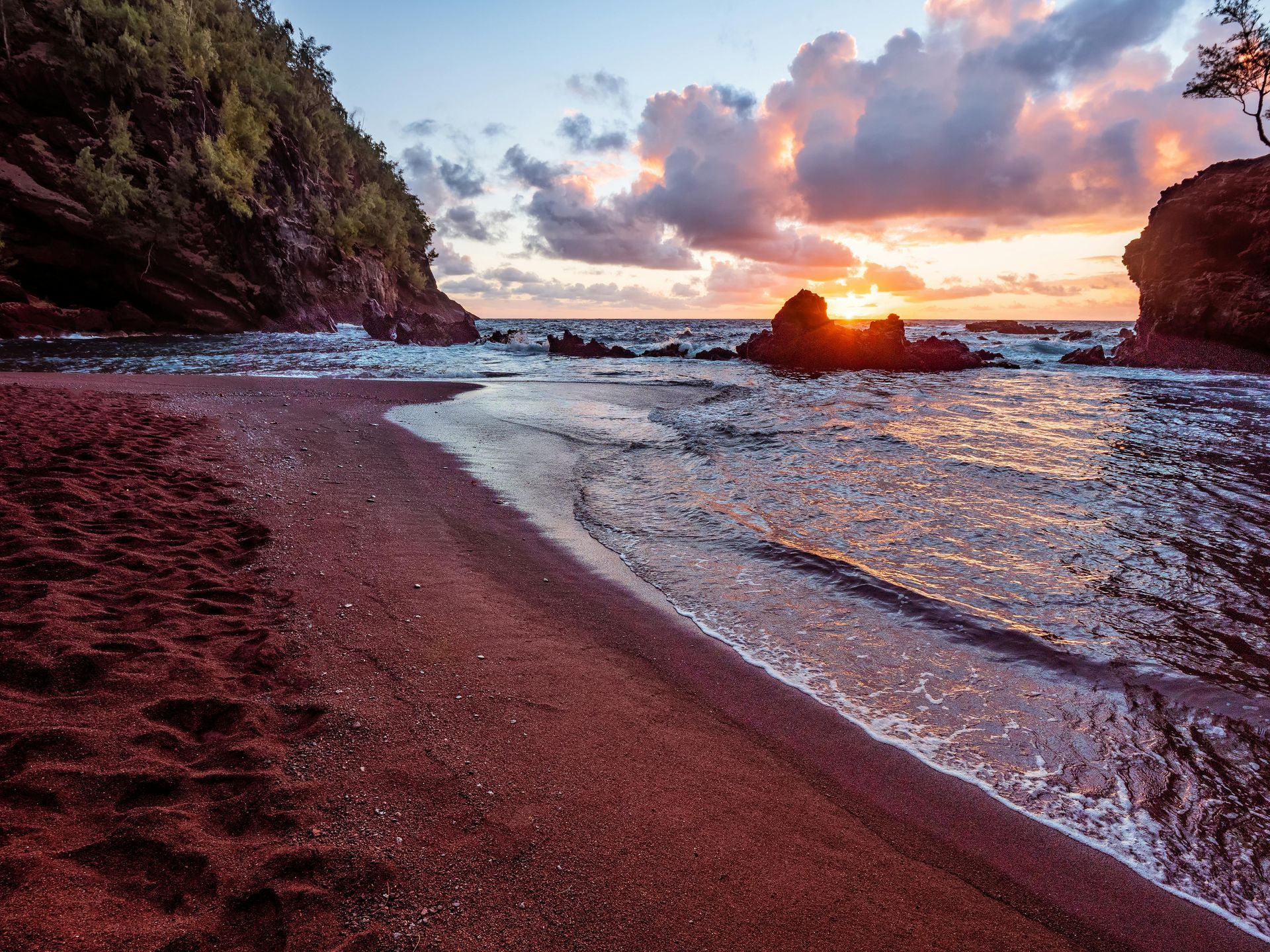 Red sand beach at sunset, waves washing ashore, rocky cliffs frame the view, fiery sky.