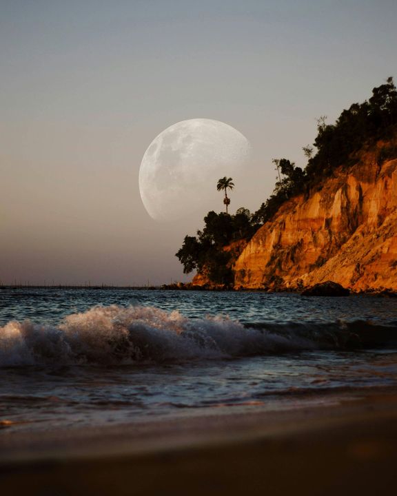 Moon rising over beach cliffs, waves crashing on shore at sunset.