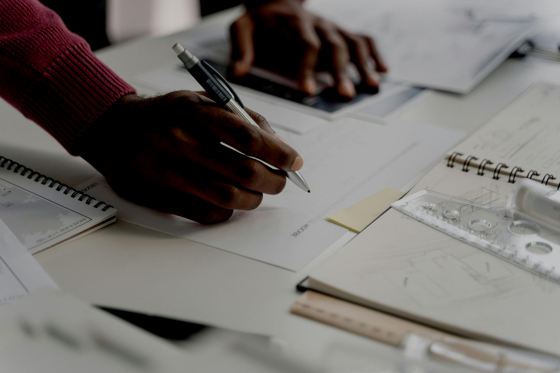 Person's hand writing with a pen on paper, with a notebook, ruler, and other papers on a desk.