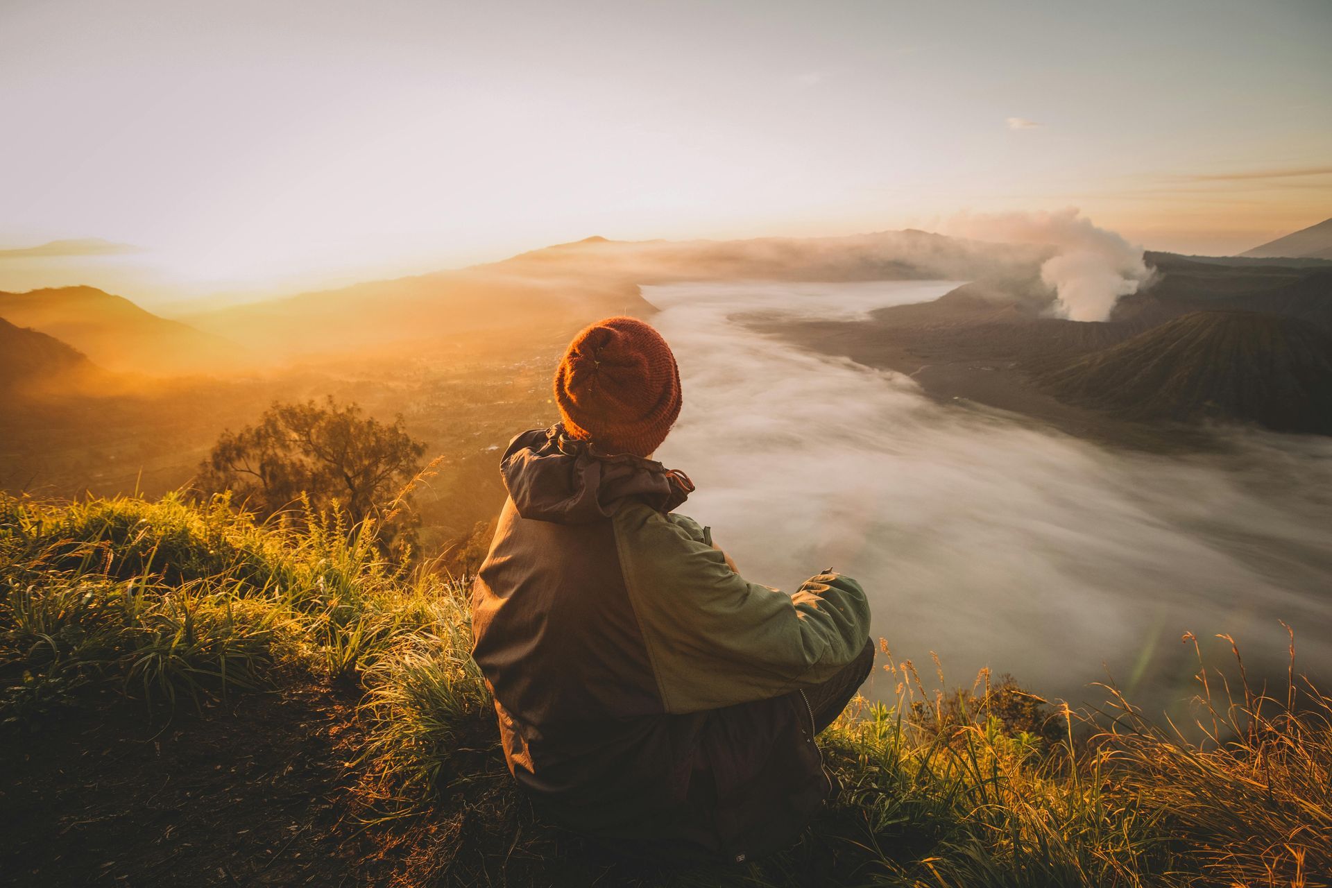 Person in orange hat sitting on a mountain peak, gazing at misty valley during sunrise.