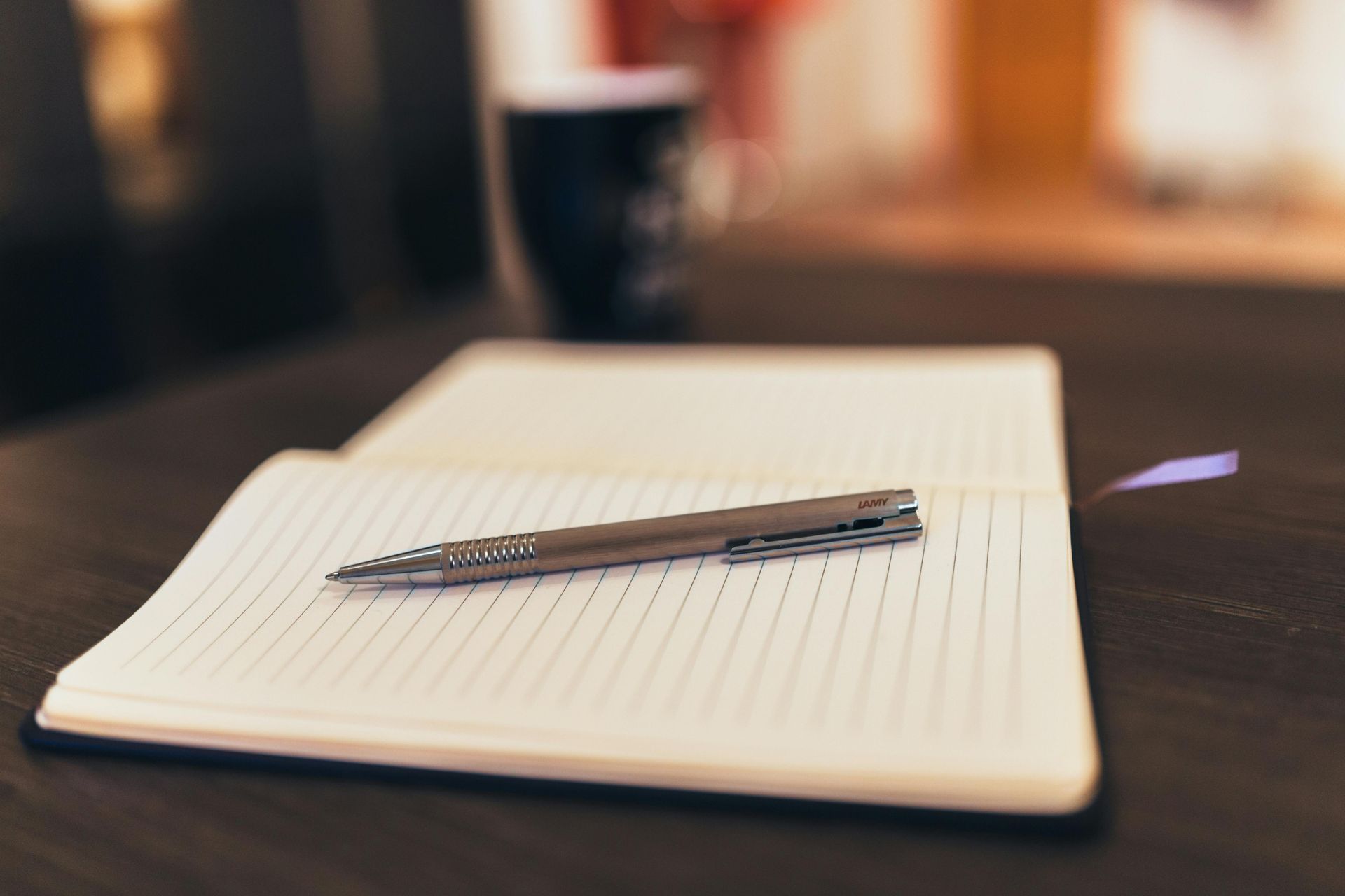 Notebook open on a dark wooden surface with a pen lying across it; background blurred with a coffee cup.