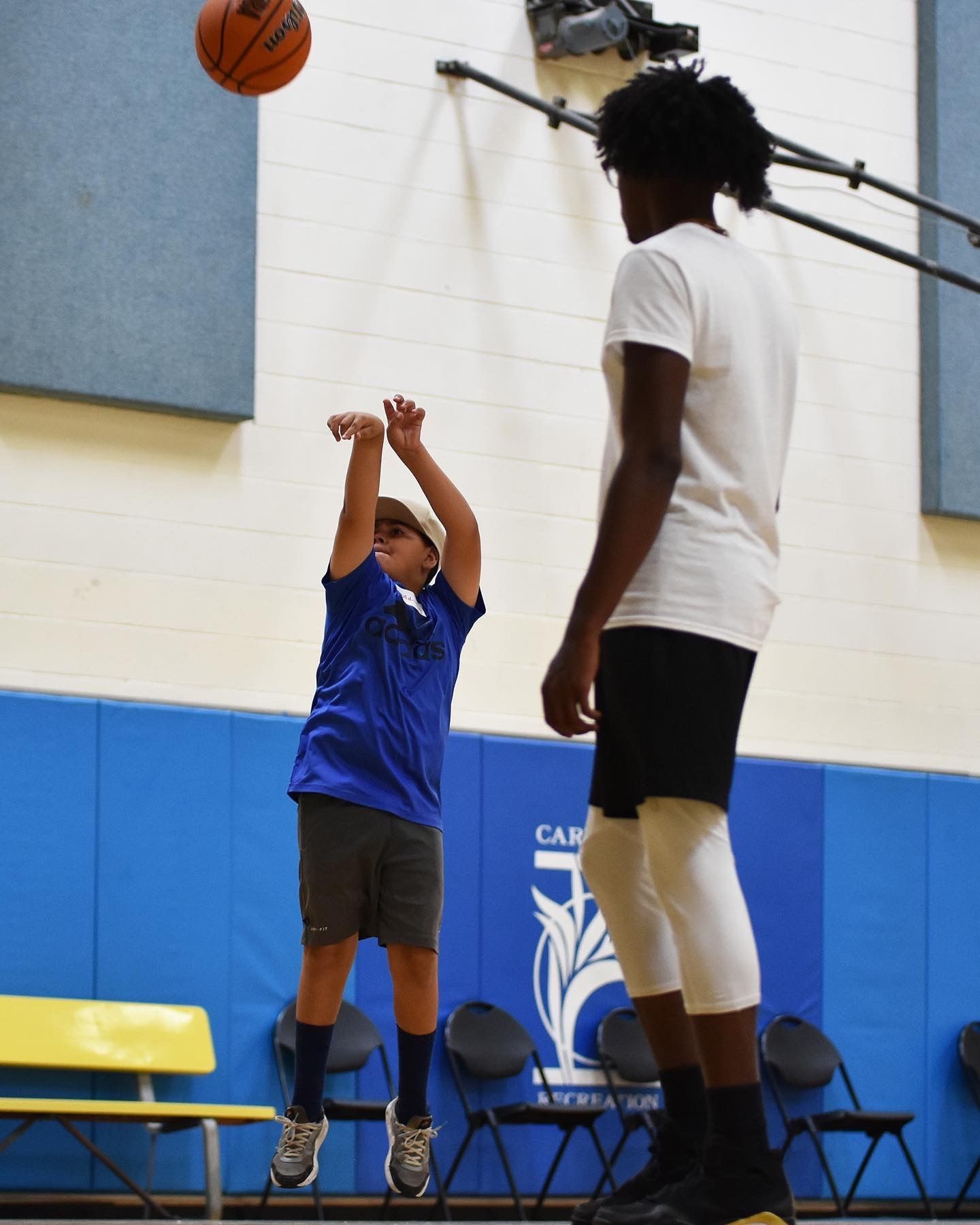 Basketball player shoots indoors while another player watches near the hoop.