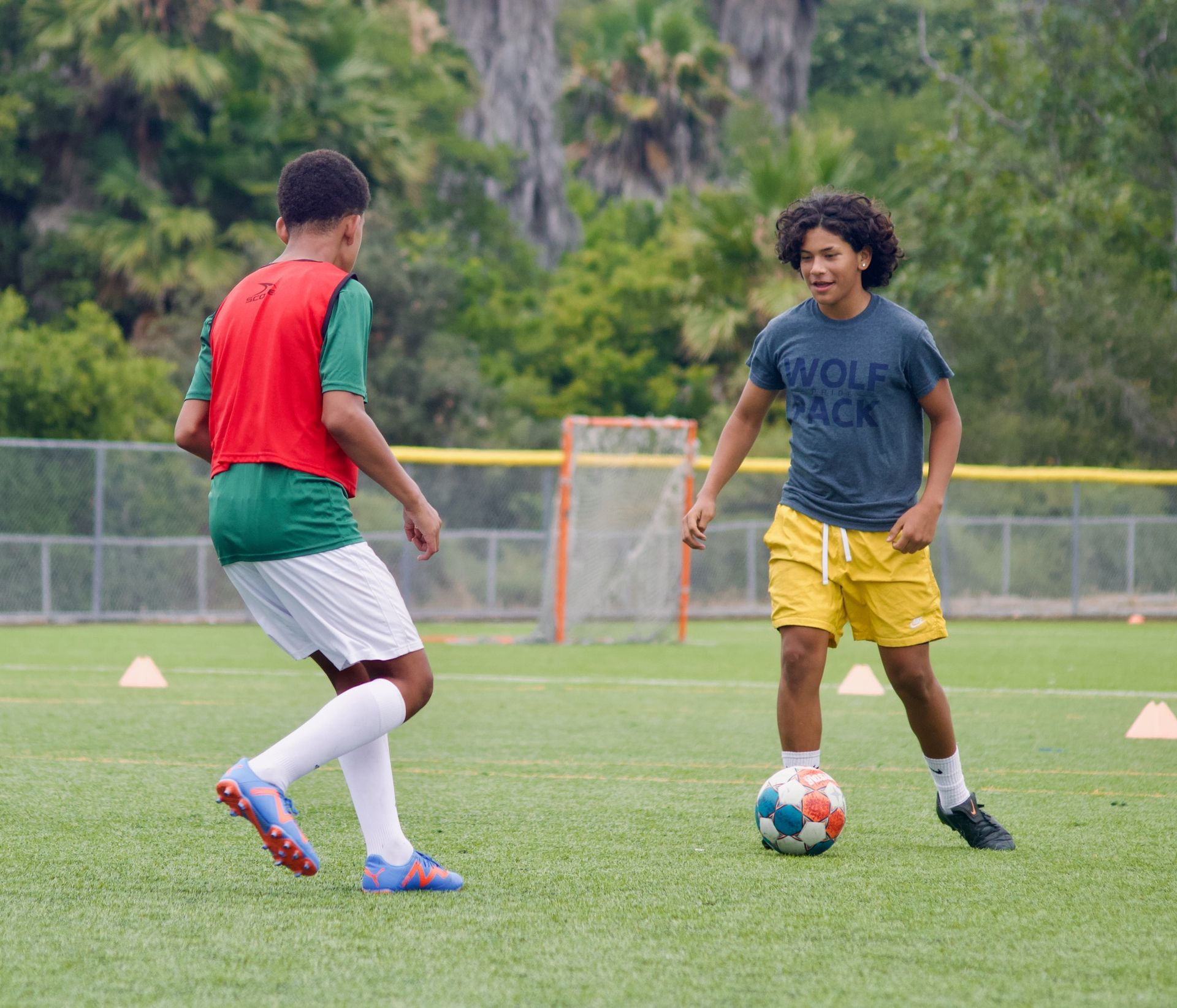 Two soccer players dribble on a grassy field, one in red and white, the other in gray and yellow.