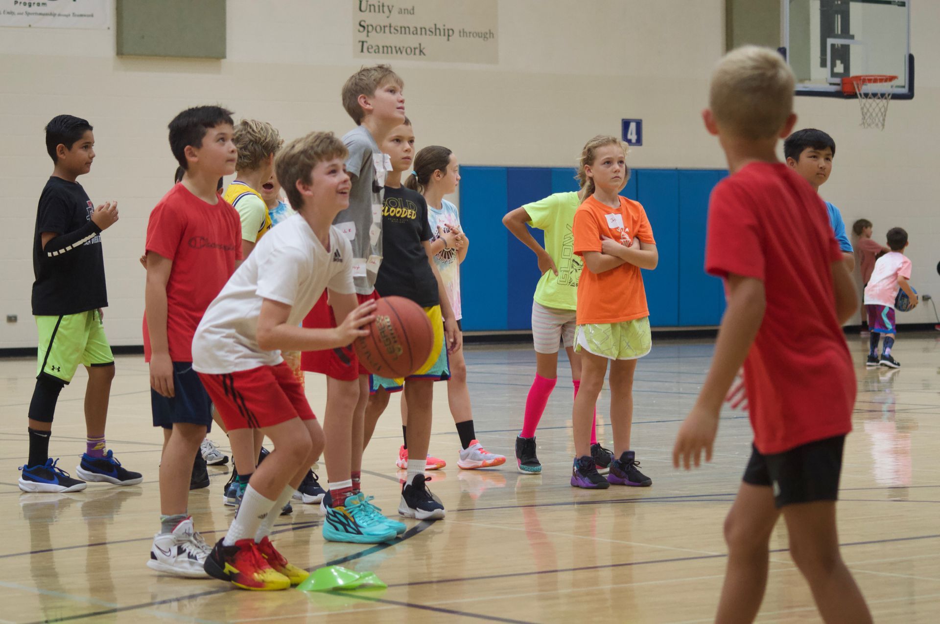 Children playing basketball in a gym, one dribbling while others watch on the court.