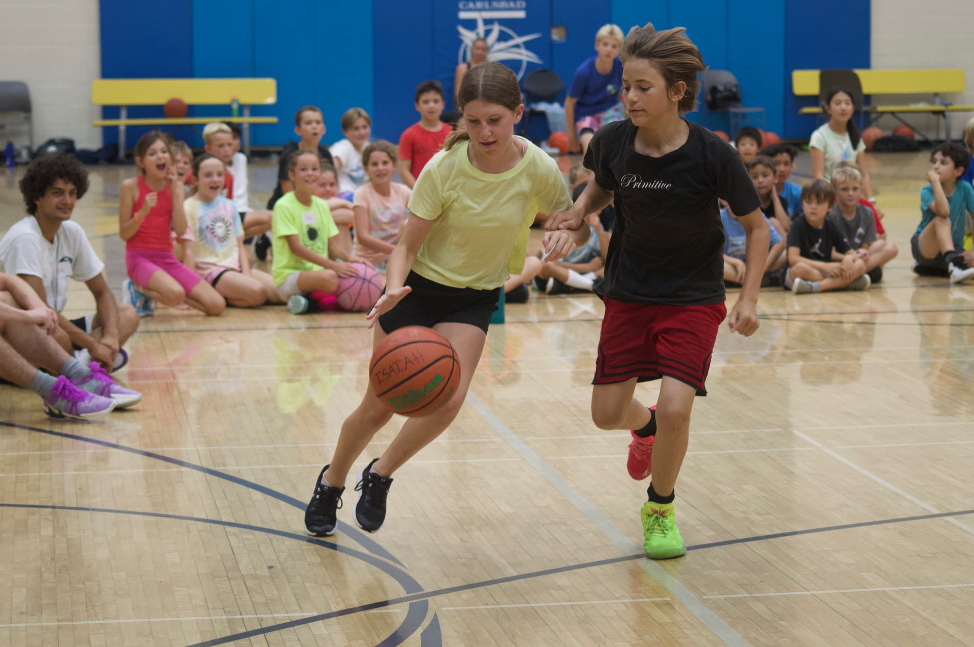 Two children playing basketball indoors, one dribbling while another defends, with spectators in the background
