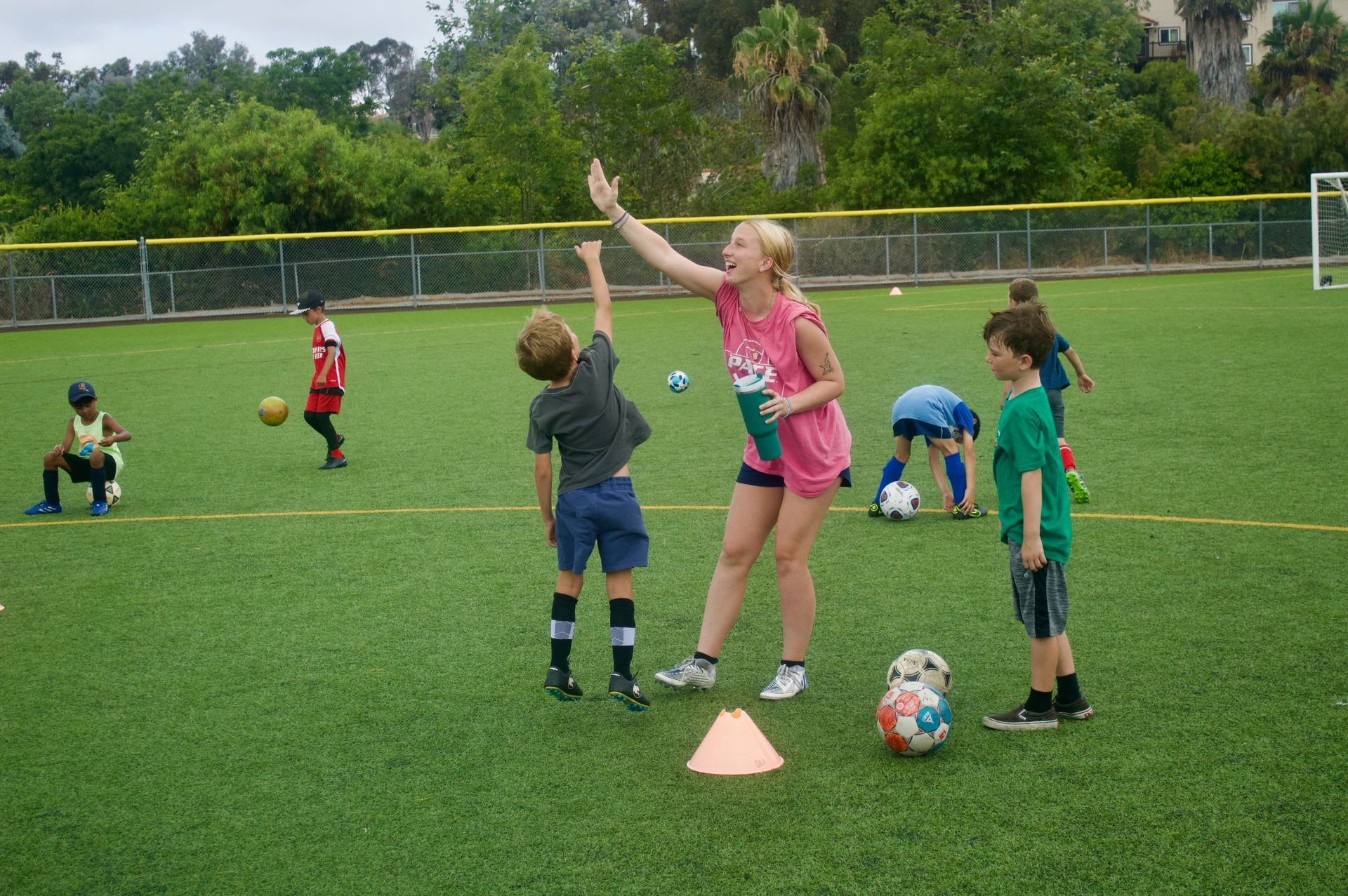 Children playing soccer drills on a grassy field, with a coach raising an arm near training cones and balls